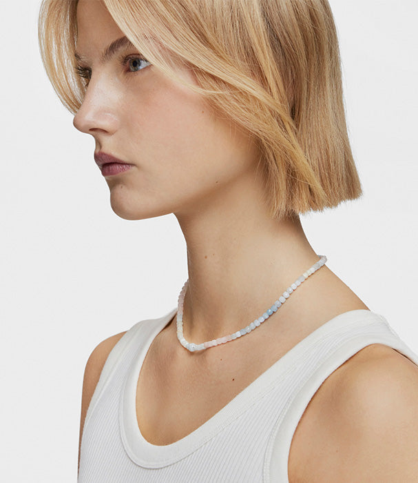 אישה עם שיער בלונד קצר עונדת שרשרת חרוזים בצבעים עדינים, חולצה לבנה ללא שרוולים