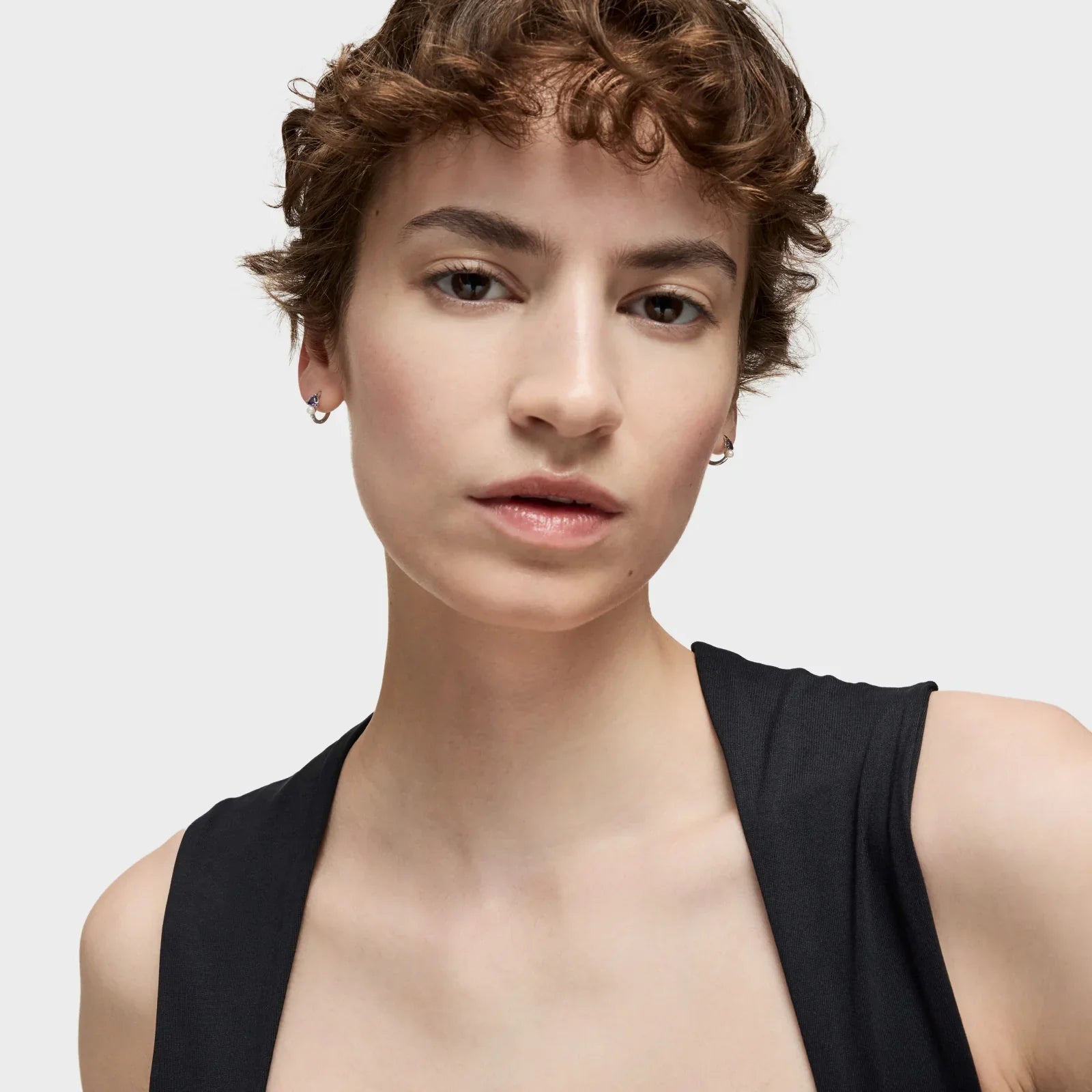 אישה עם שיער קצר מתולתל עונדת עגילים, לובשת גופיה שחורה, רקע לבן, צילום CHOZEN