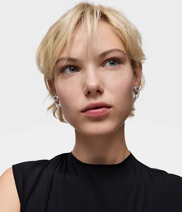 דוגמנית עם שיער בלונדיני קצר, עונדת עגילים כסופים מבריקים, לובשת חולצה שחורה, רקע בהיר. CHOZEN