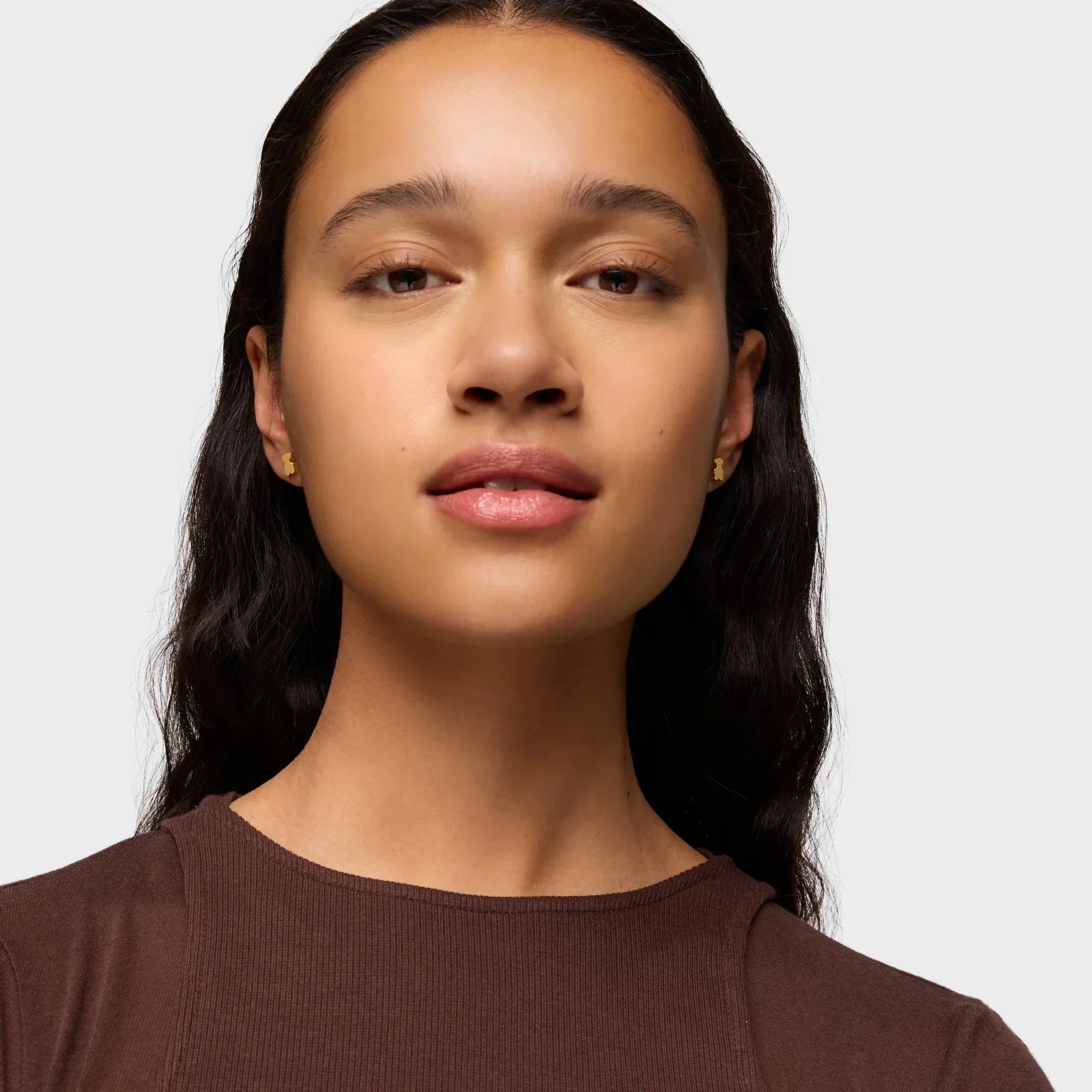 אישה עם עגילי זהב עדינים, שיער כהה גלי וחולצה חומה, רקע לבן, קמפיין מותגים CHOZEN