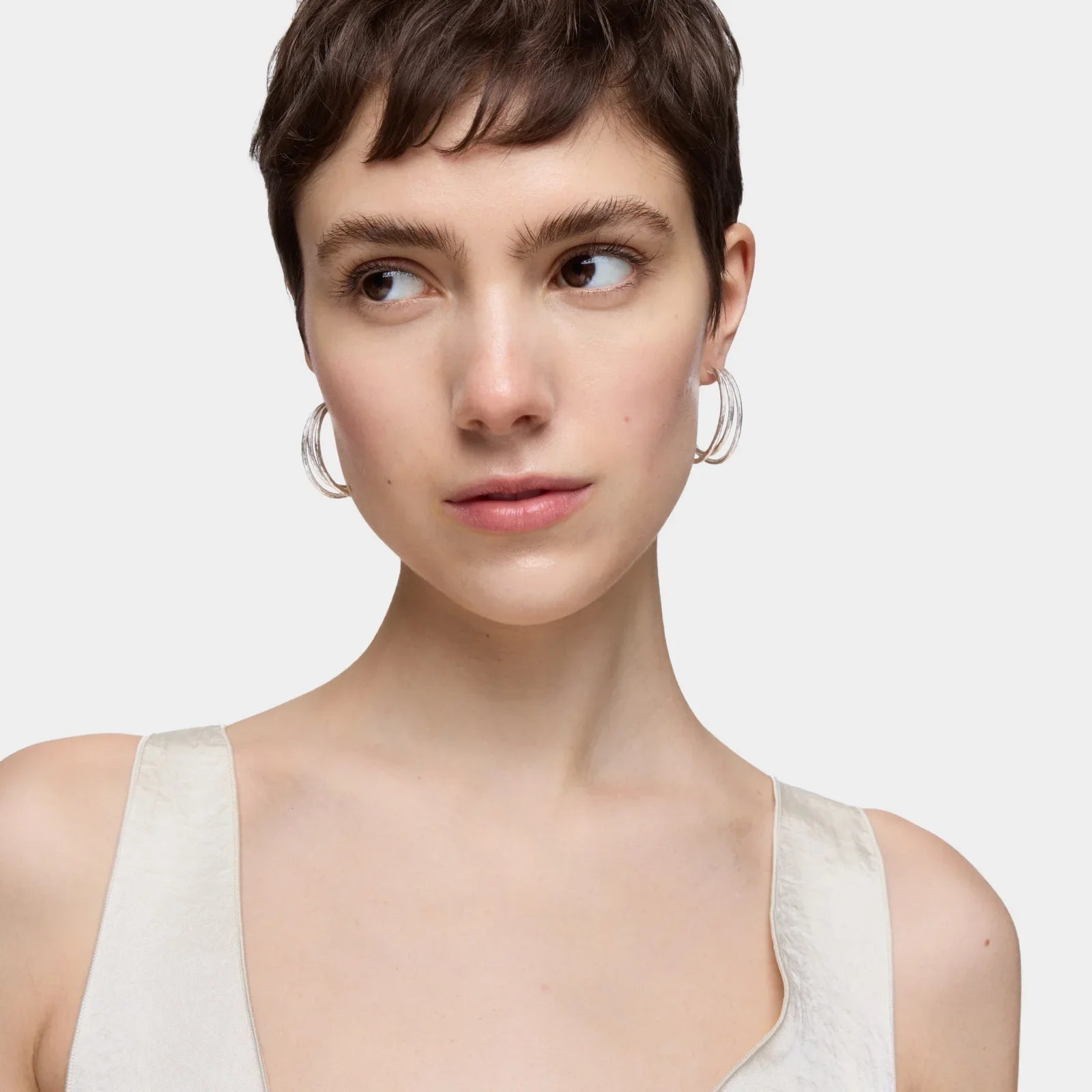 אישה עם עגילי חישוק כסופים, שיער קצר ולבוש בהיר, צולם על רקע לבן ל-CHOZEN