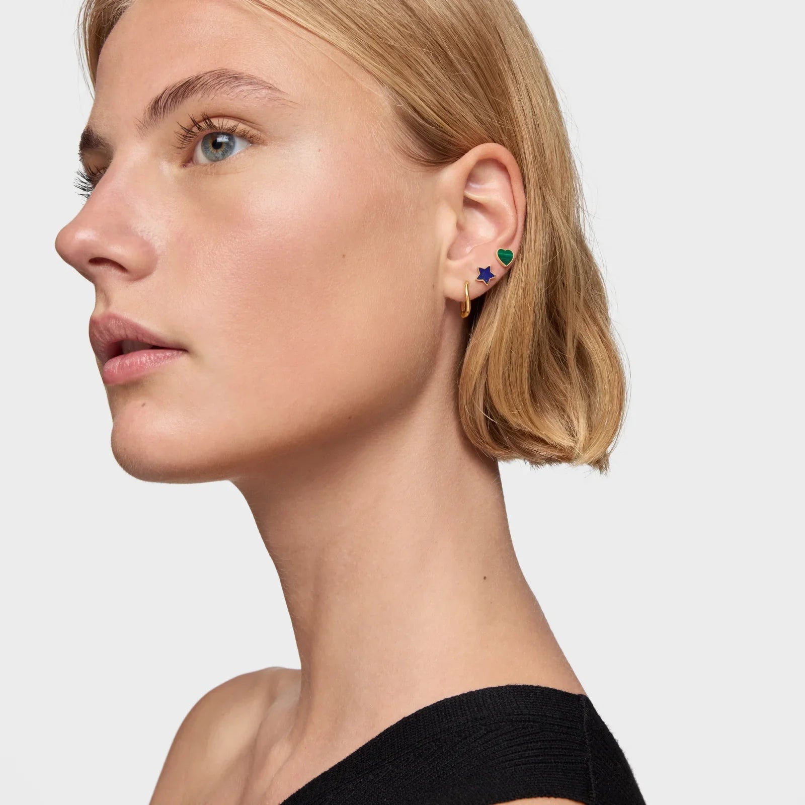 אישה עם שיער בלונדיני קצר עונדת שלושה עגילים צבעוניים, דוגמת כוכב ולב, רקע בהיר, CHOZEN תכשיטים