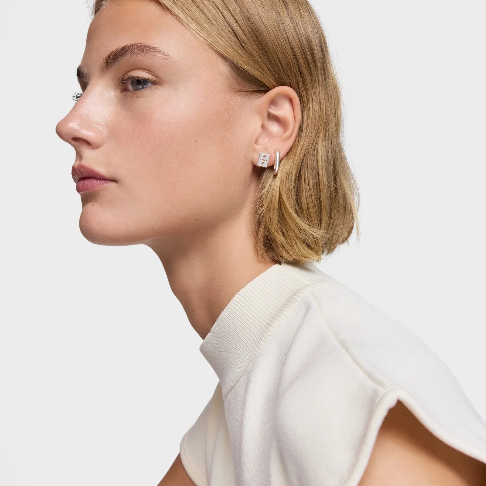 אישה עם שיער בלונדיני קצר, עונדת עגילי כסף מודרניים, חולצה לבנה, רקע לבן, CHOZEN מותגים מובילים