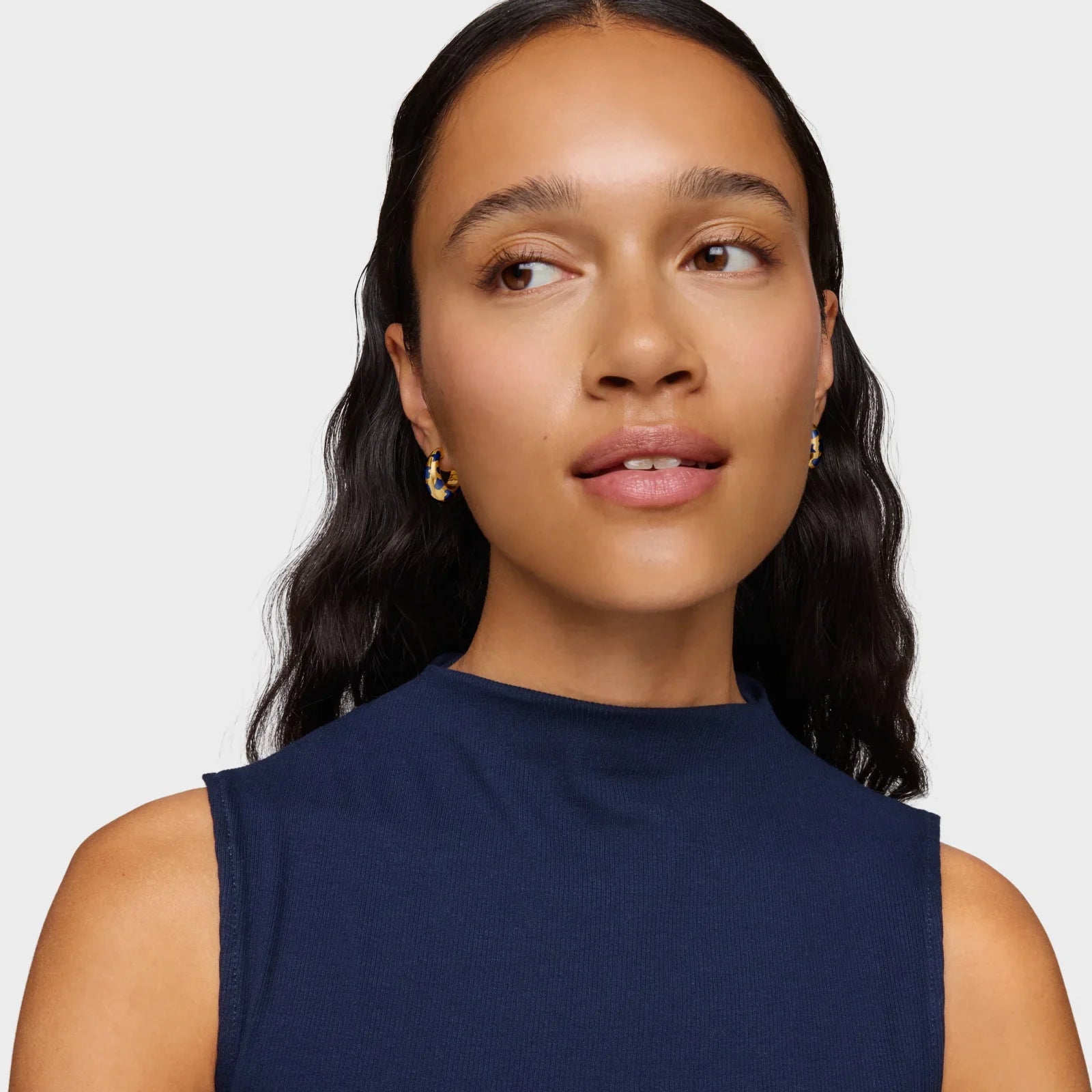 אישה עם שיער כהה בגלים, עונדת עגילי זהב יוקרתיים, גופיה כחולה, צילום קמפיין CHOZEN