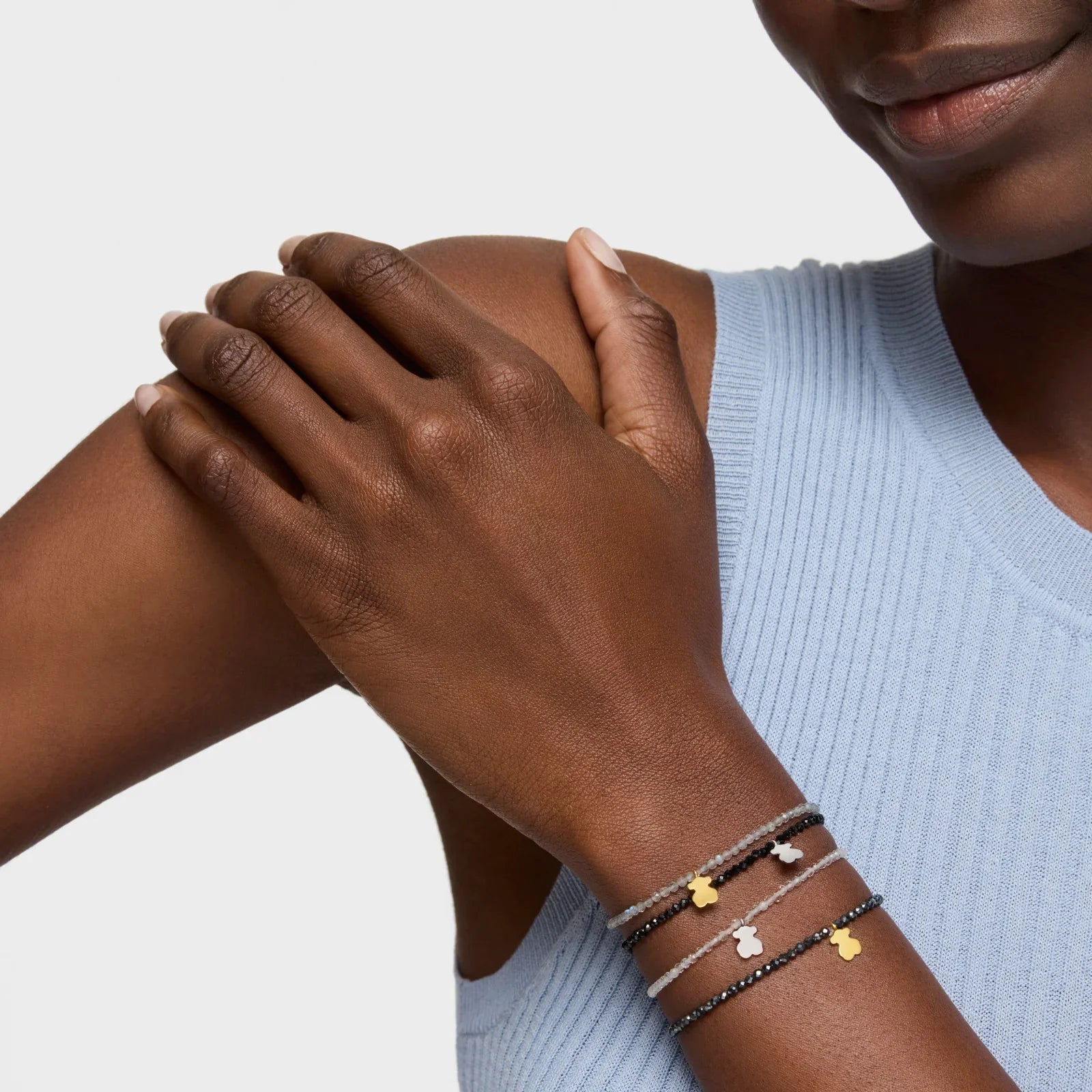 יד אישה עם שלושה צמידי חרוזים דקים ותליוני דובי בצבעים זהב וכסף, חולצת סריג תכלת, צ׳וזן מותגים מובילים