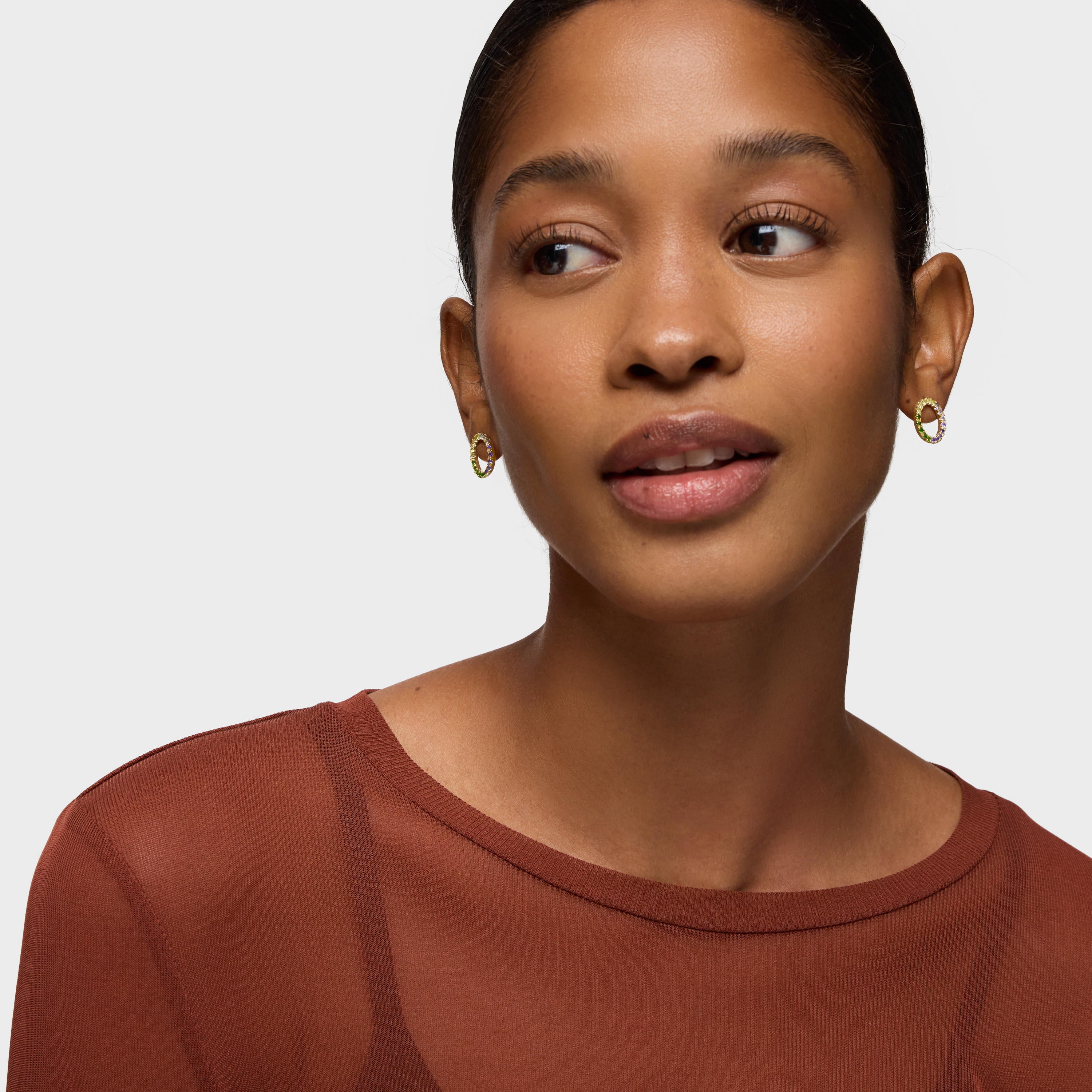 אישה עונדת עגילי חישוק זהב עם אבנים צבעוניות, חולצה חומה, רקע בהיר