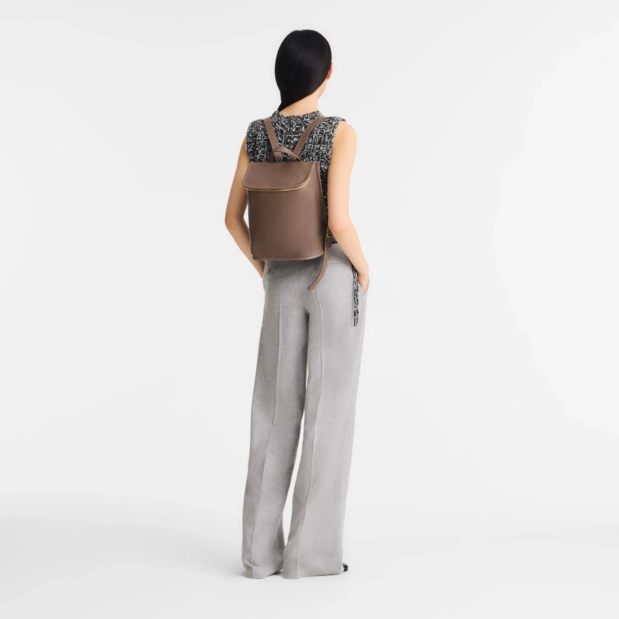 אישה עם תיק גב חום מבית CHOZEN, לובשת מכנסיים אפורים וחולצה שחורה לבנה, רקע לבן