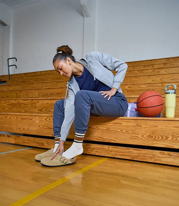 אישה יושבת על ספסל במגרש כדורסל, נועלת כפכפי Birkenstock, כדורסל ובקבוק לידה