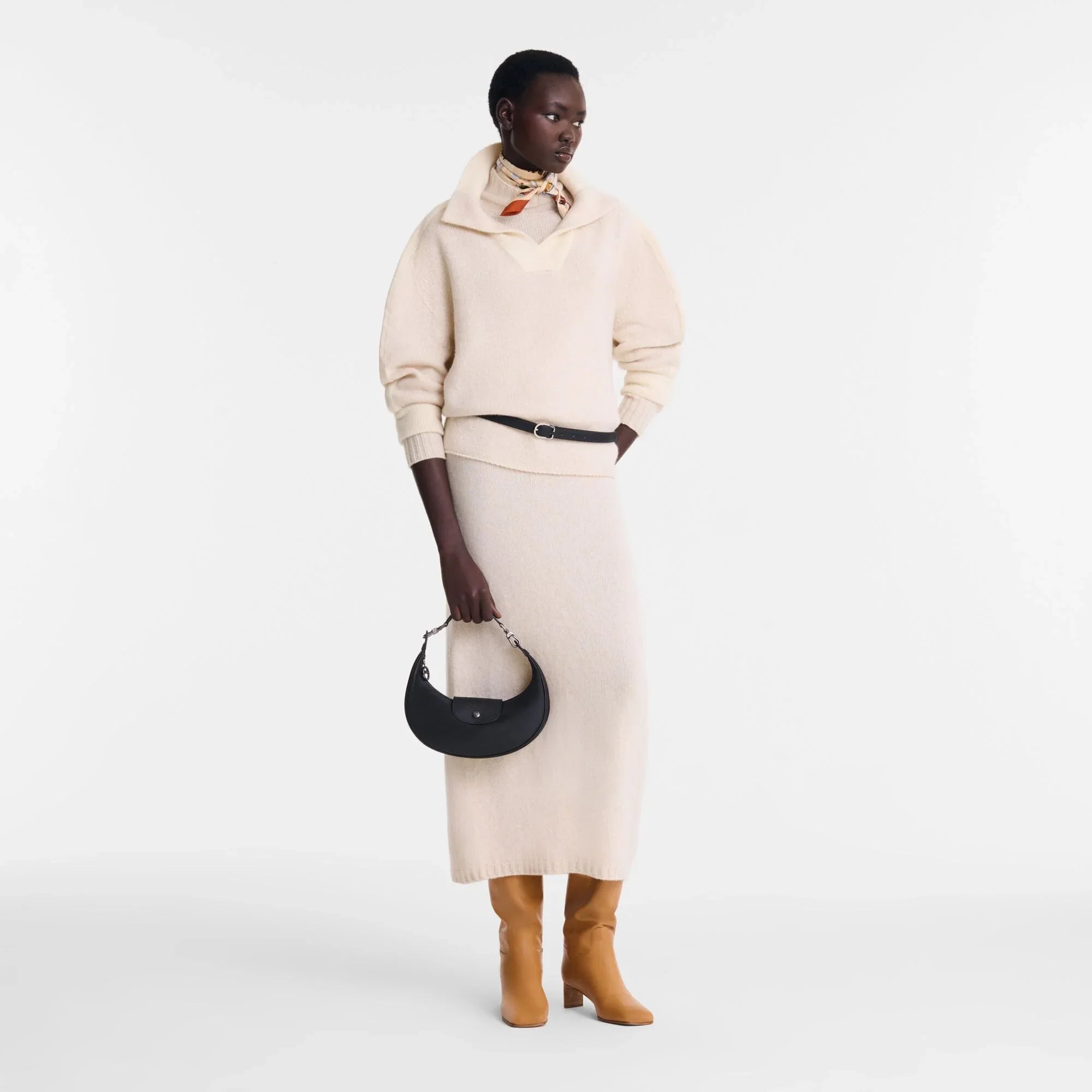 דוגמנית לובשת סוודר וחצאית שמנת, מחזיקה תיק שחור, מגפיים חומים מבית CHOZEN