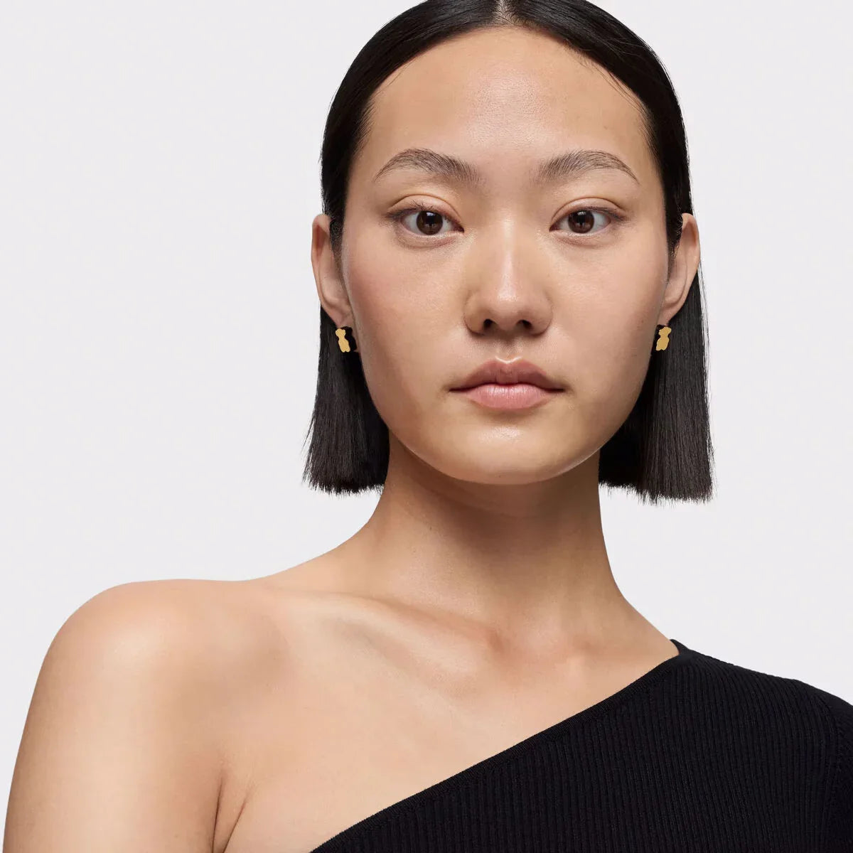 אישה עם שיער קצר עונד עגילי זהב, לבושה בטופ שחור אסימטרי, רקע בהיר, CHOZEN