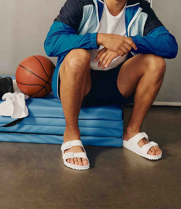 גבר יושב עם סנדלים לבנים, בגדי ספורט, כדורסל ומזרנים כחולים בחדר כושר