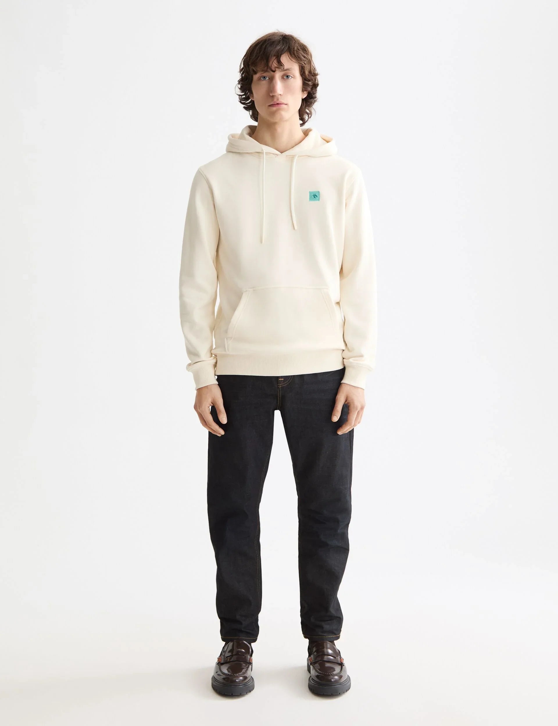 גבר לובש קפוצ'ון בז' עם לוגו, ג'ינס כהה ונעליים שחורות על רקע לבן, אופנת CHOZEN