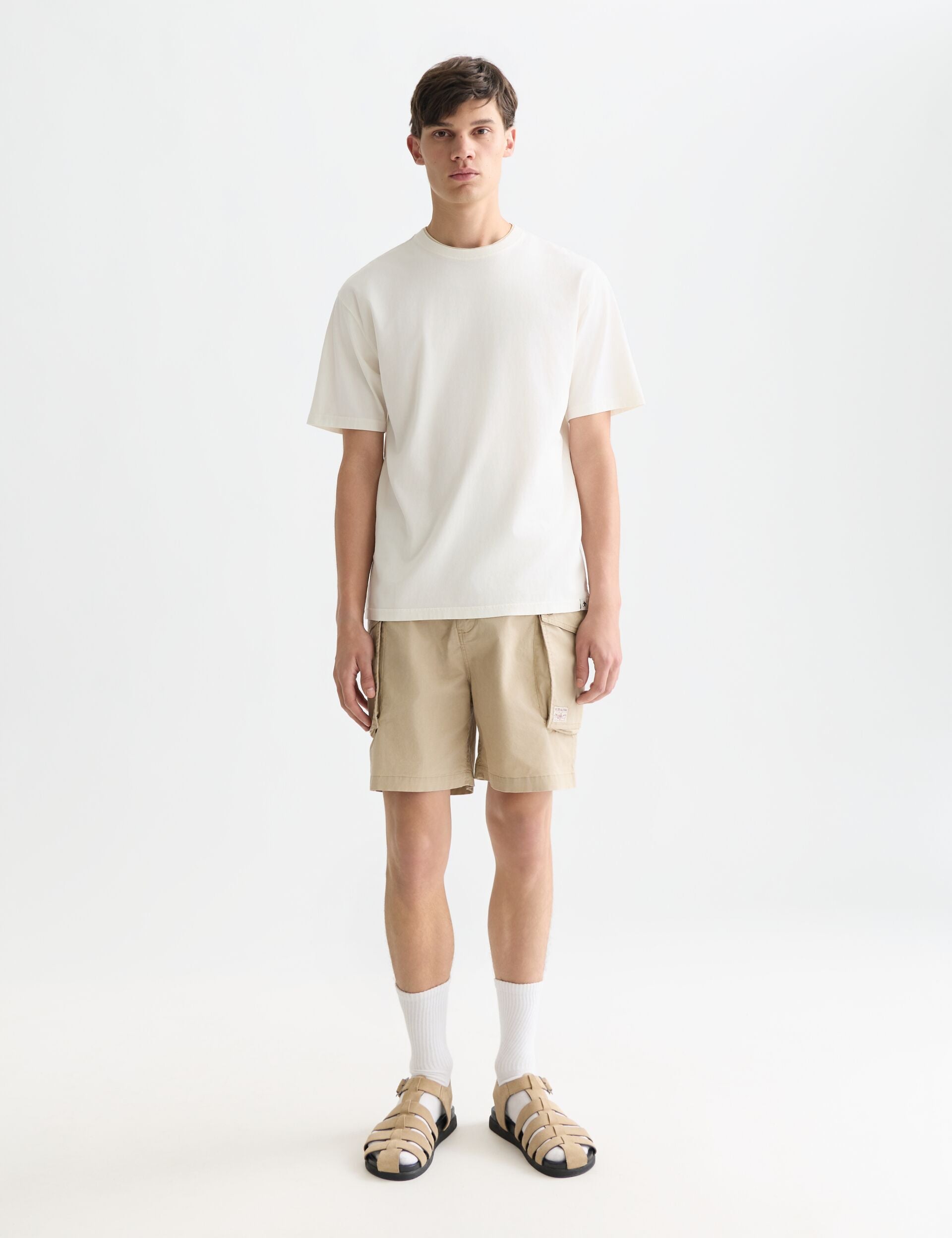 גבר צעיר לובש חולצה לבנה, שורטס בז', סנדלים וגרביים לבנות, רקע סטודיו לבן, CHOZEN