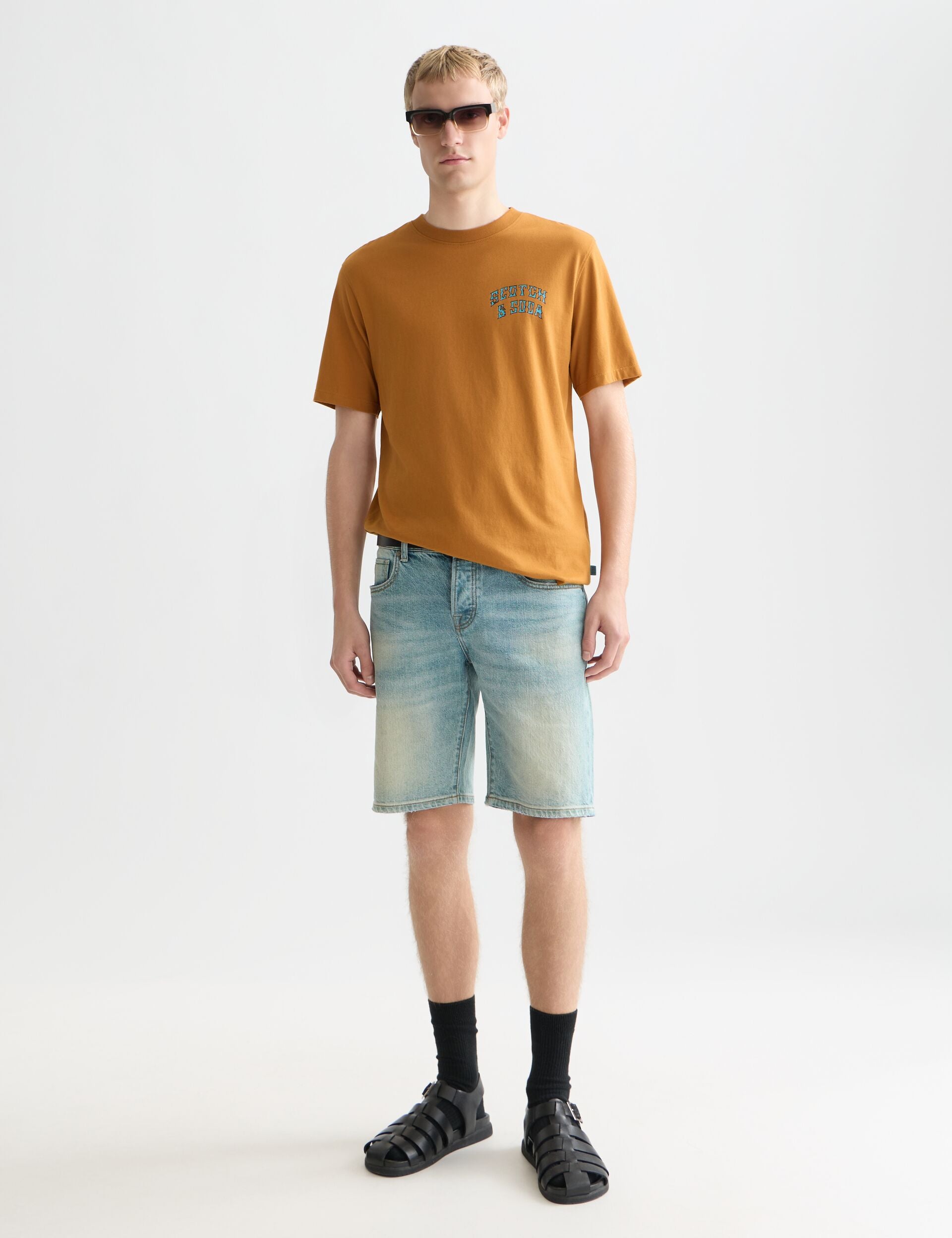 דגם גבר עם חולצה כתומה של CHOZEN, מכנסי ג'ינס קצרים, סנדלים שחורים ומשקפי שמש