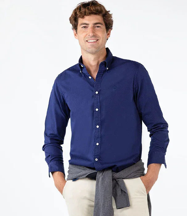 גבר עם חולצה כחולה מכופתרת של מותג CHOZEN, לובש מכנסי בז' וקשור סוודר אפור במותניים