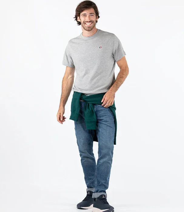 גבר מחייך לובש חולצת טי אפורה, ג'ינס ונעלי ספורט, עומד על רקע לבן. CHOZEN מותגי אופנה.