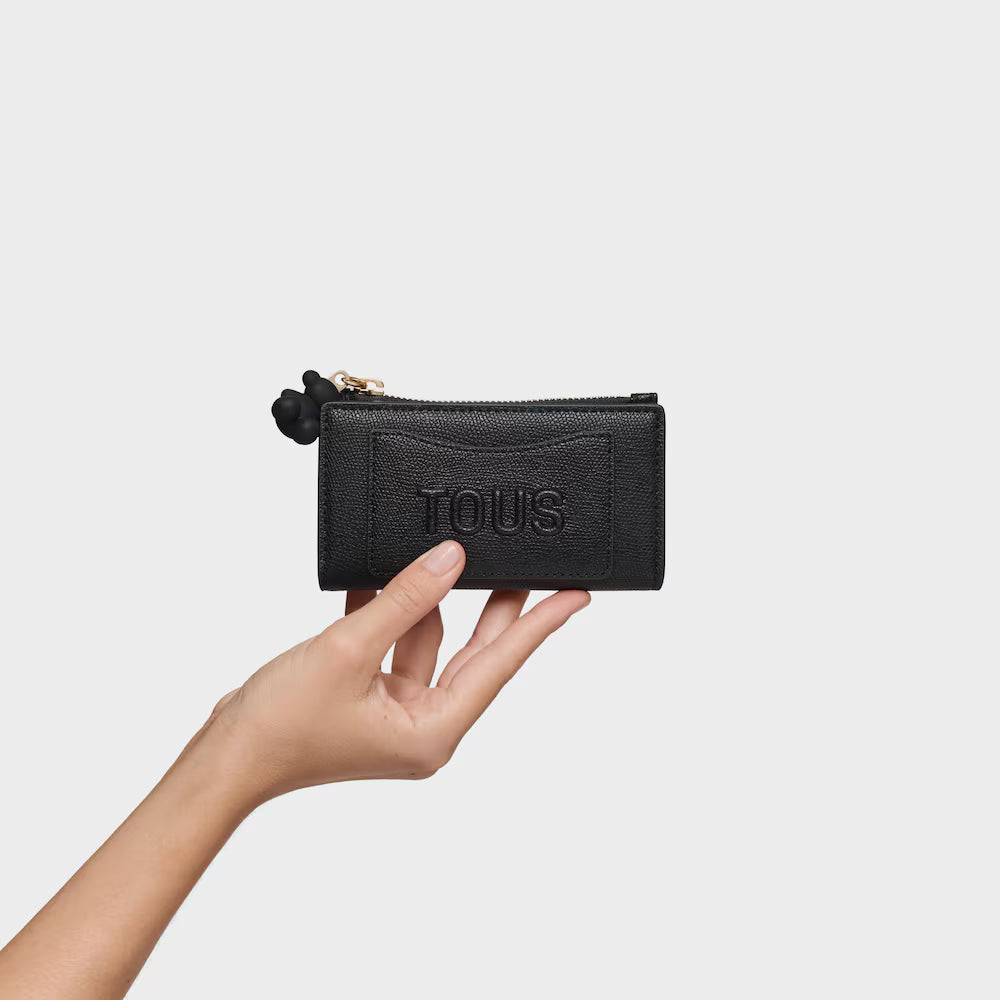ארנק עור שחור קטן עם רוכסן, מוחזק ביד, רקע לבן, לוגו TOUS בחזית