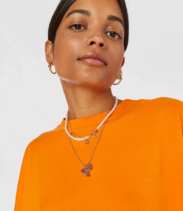 אישה עם חולצת טי כתומה, עונדת תכשיטי זהב ושרשרת פנינים, אופנה מבית CHOZEN