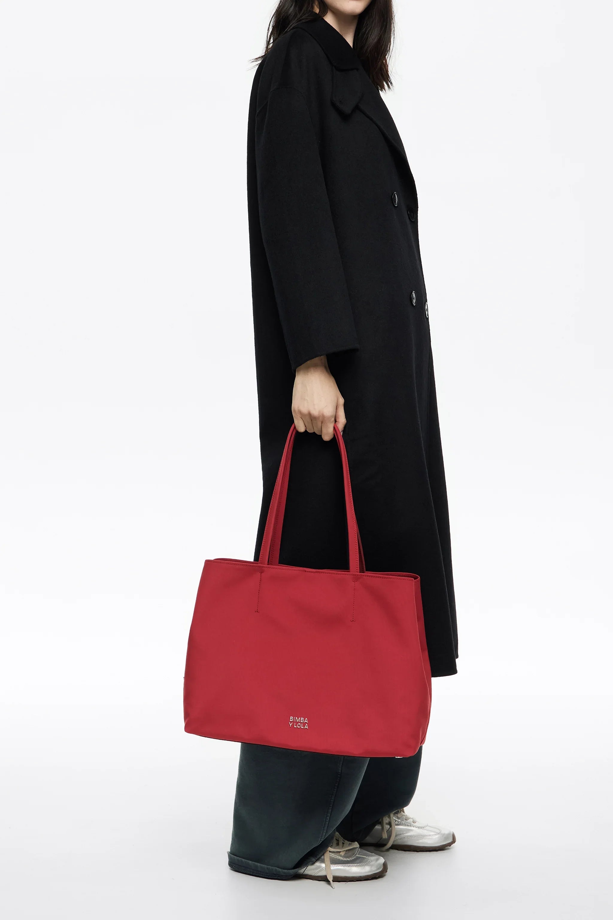 אישה במעיל שחור מחזיקה תיק יד אדום מבית BIMBA Y LOLA על רקע לבן, CHOZEN מותגים מובילים