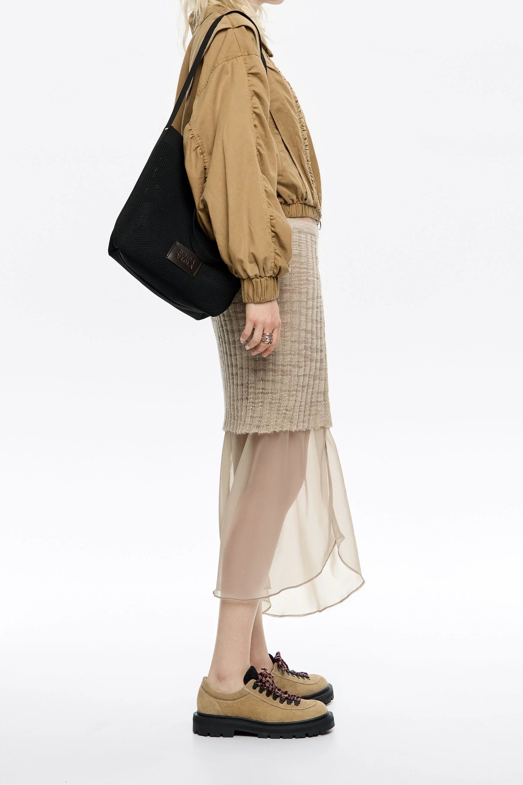 אישה עם חצאית אופנתית מבד שקוף, ג’קט חום, נעלי סניקרס בז’ ותיק כתף שחור מחנות המותגים CHOZEN