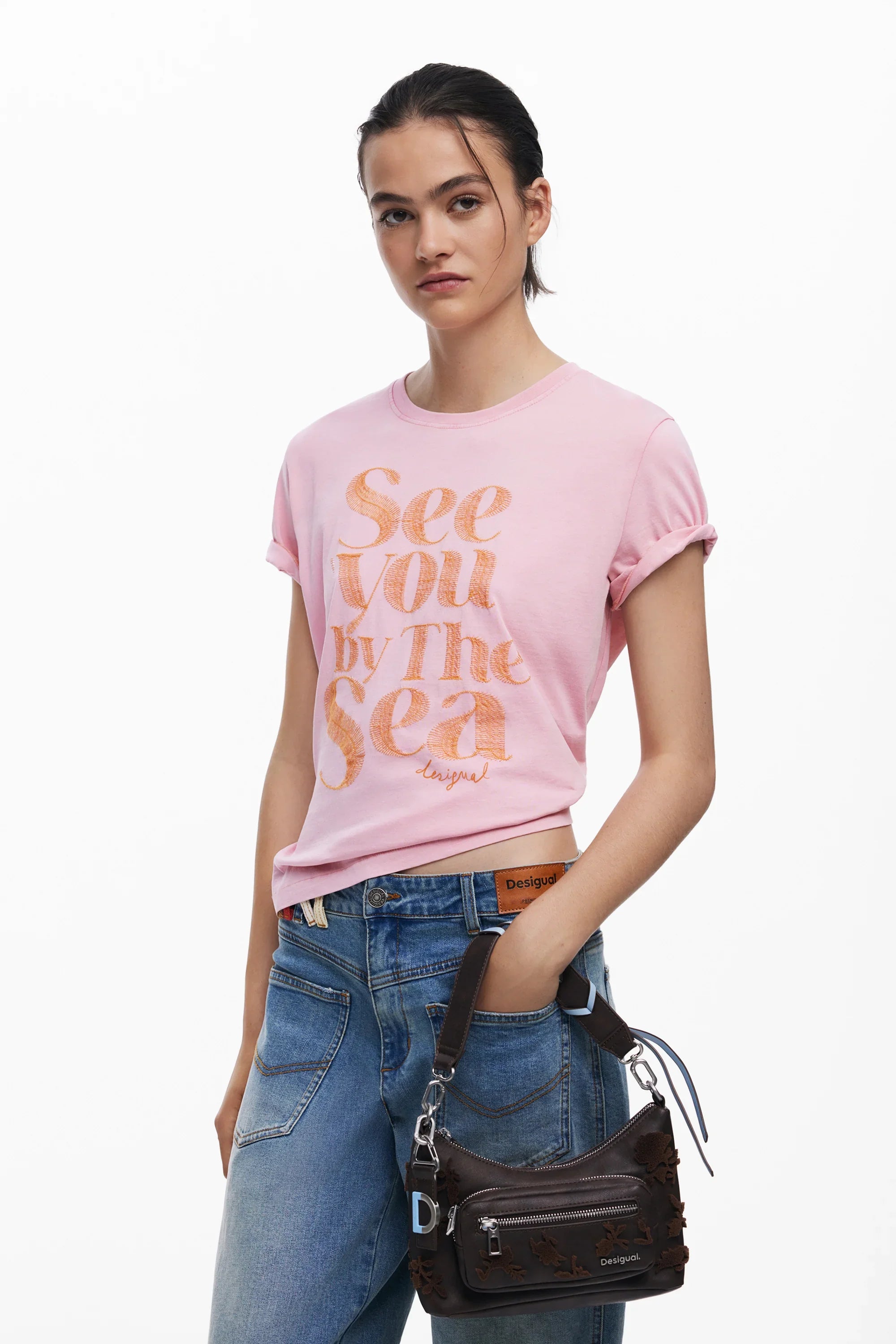 אישה עם חולצת טי ורודה של Desigual עם כיתוב, ג'ינס ותיק יד חום, מותגים מובילים בחנות CHOZEN