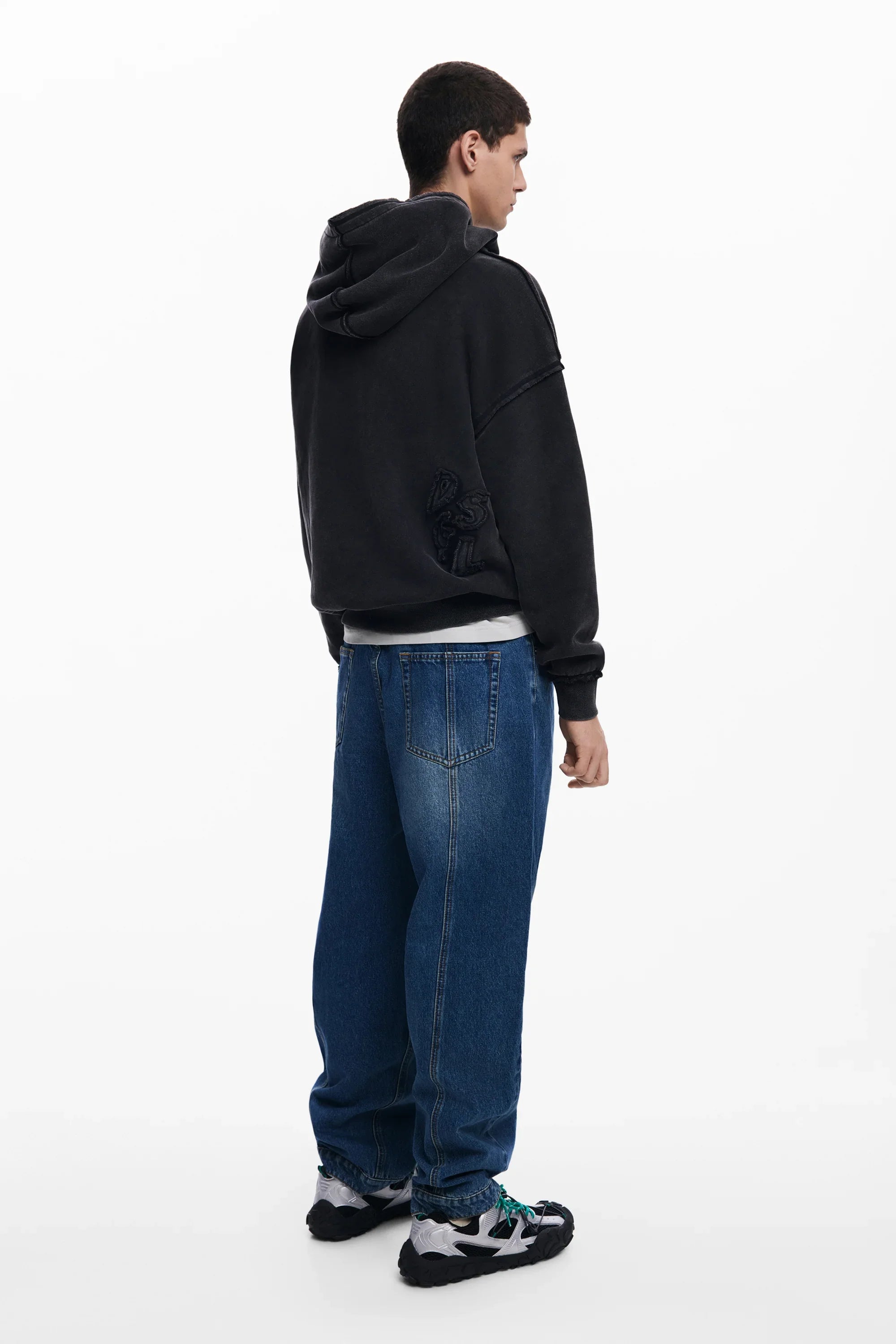 גבר לובש קפוצ'ון שחור של CHOZEN, ג'ינס כחול ונעלי סניקרס, על רקע לבן נקי