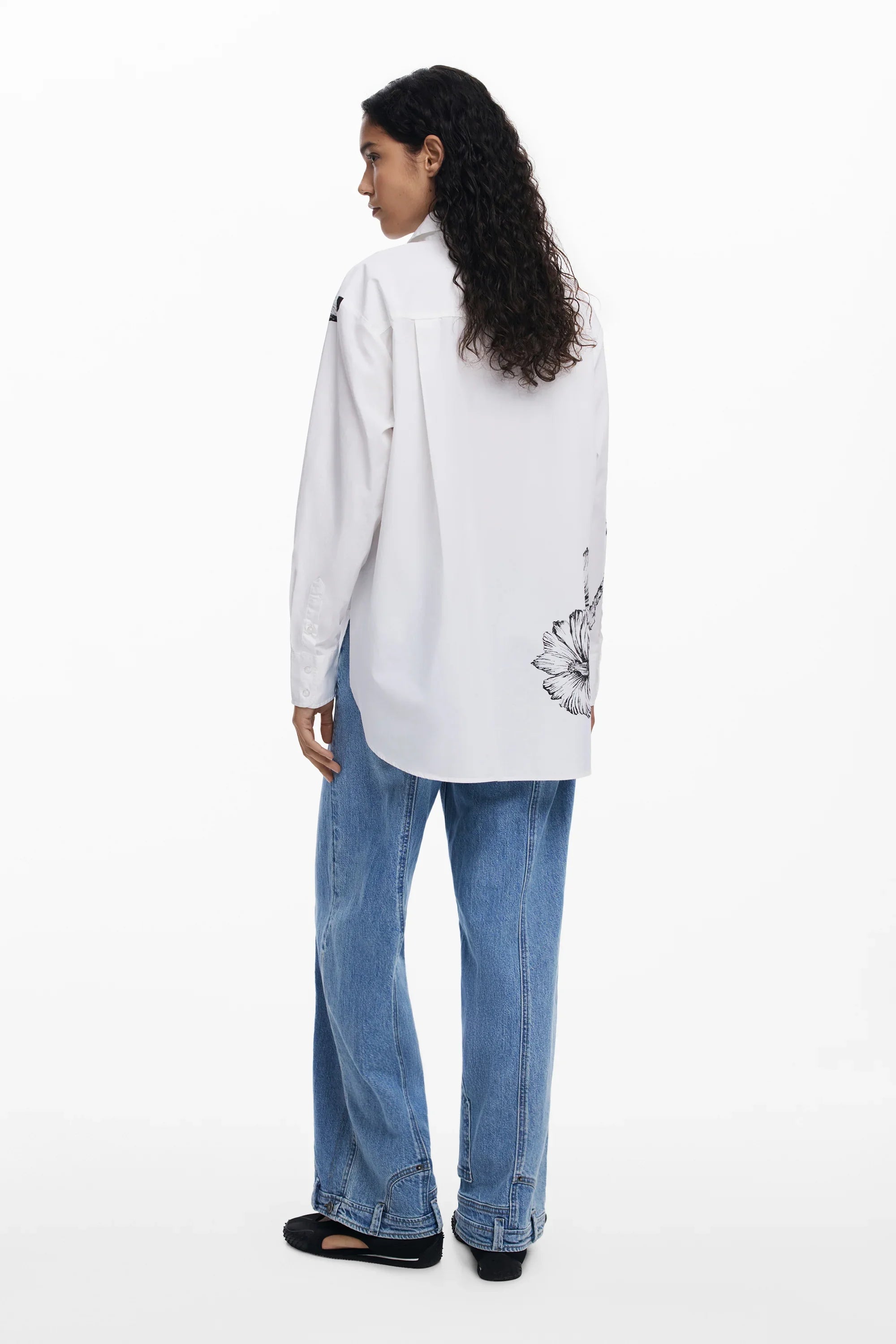 אישה לובשת חולצה לבנה עם ציור פרח בגב, ג'ינס כחול ונעליים שחורות, אופנת CHOZEN