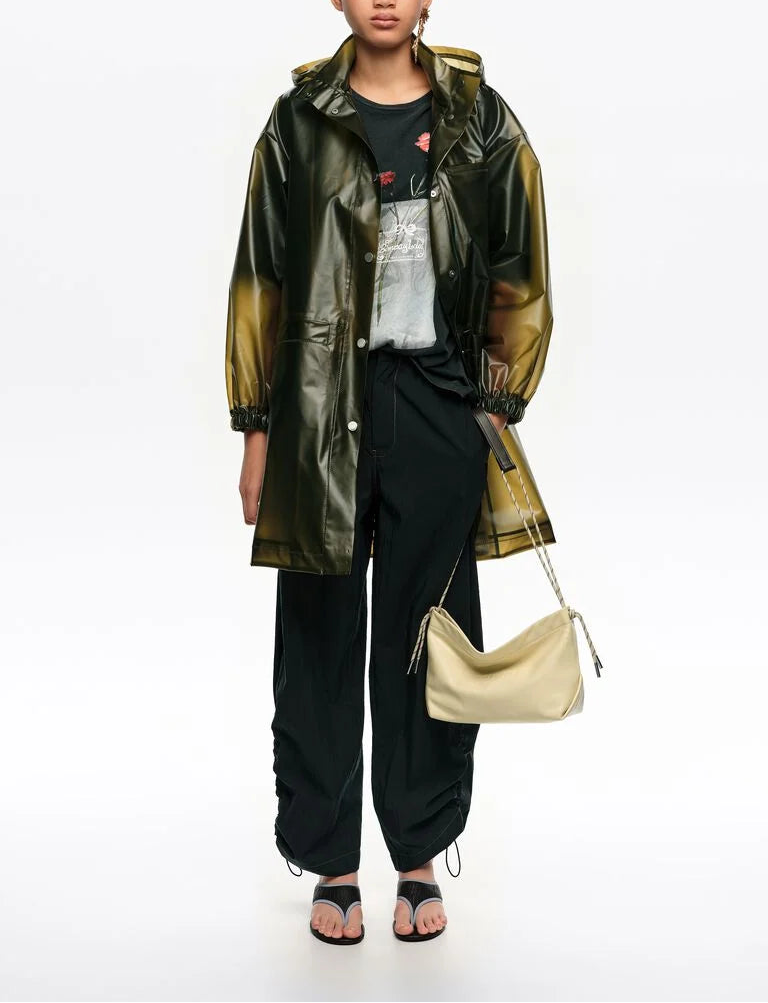 אישה לובשת מעיל גשם ירוק שקוף, מכנסיים שחורים, חולצה כהה ותיק כתף בהיר