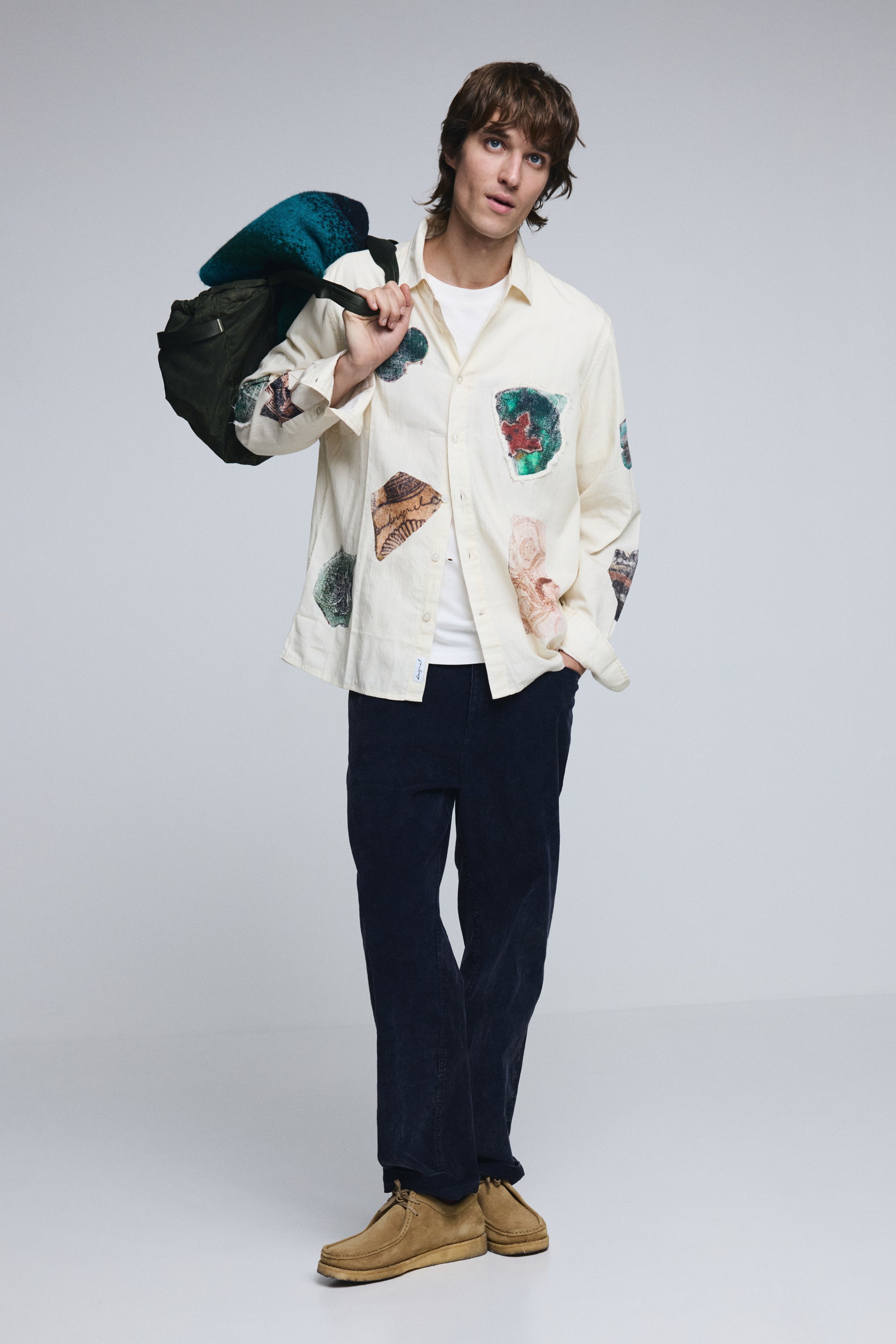 גבר עם חולצה לבנה מודפסת בטקסטורות צבעוניות, מכנס כהה ונעליים בהירות, מחזיק מעיל כתף, רקע סטודיו לבן