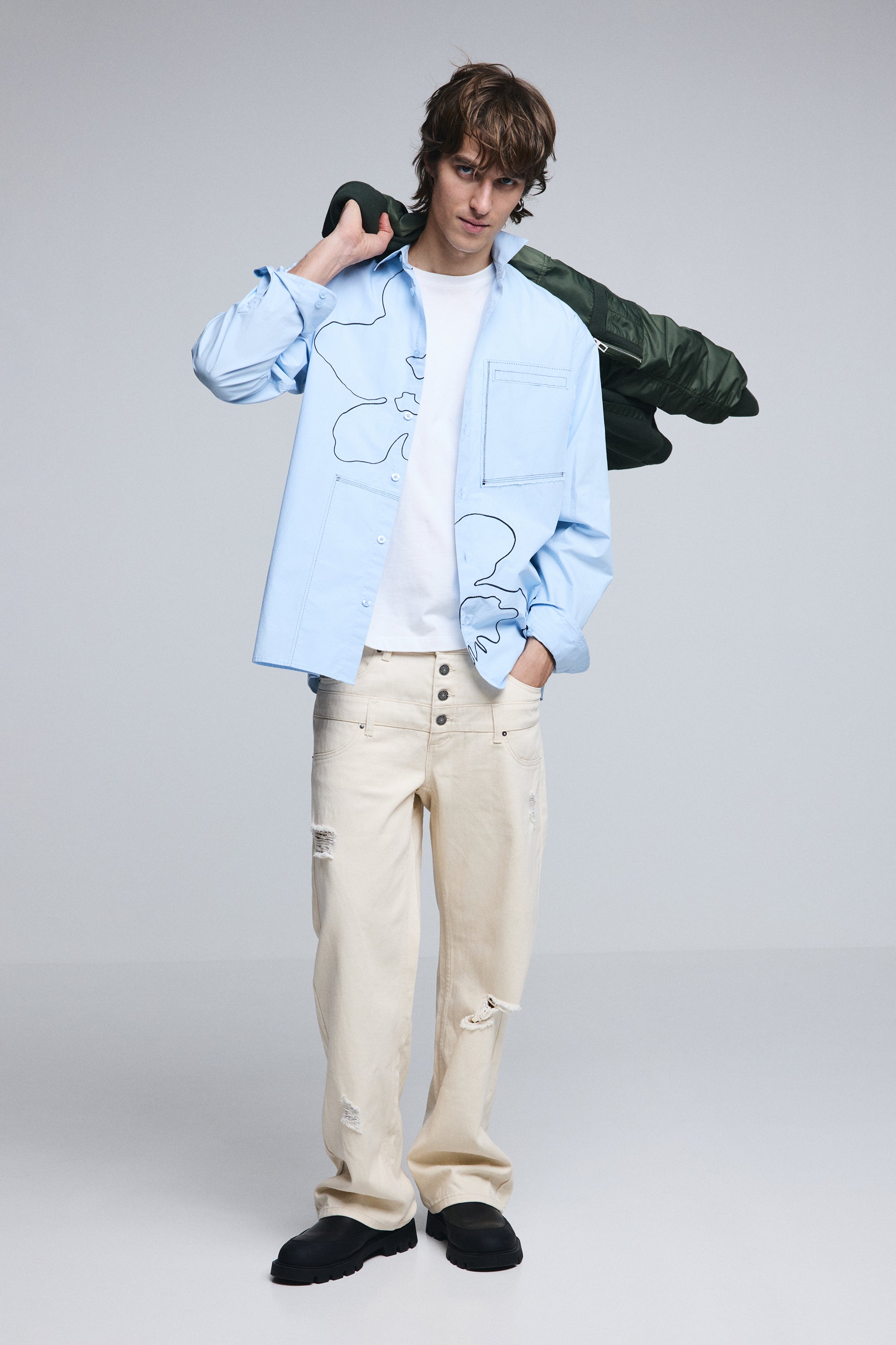 דוגמן לובש חולצה כחולה עם הדפס קווי מתאר, ג׳ינס בז׳ בגזרה רחבה וקרעים ונעלי שחור.