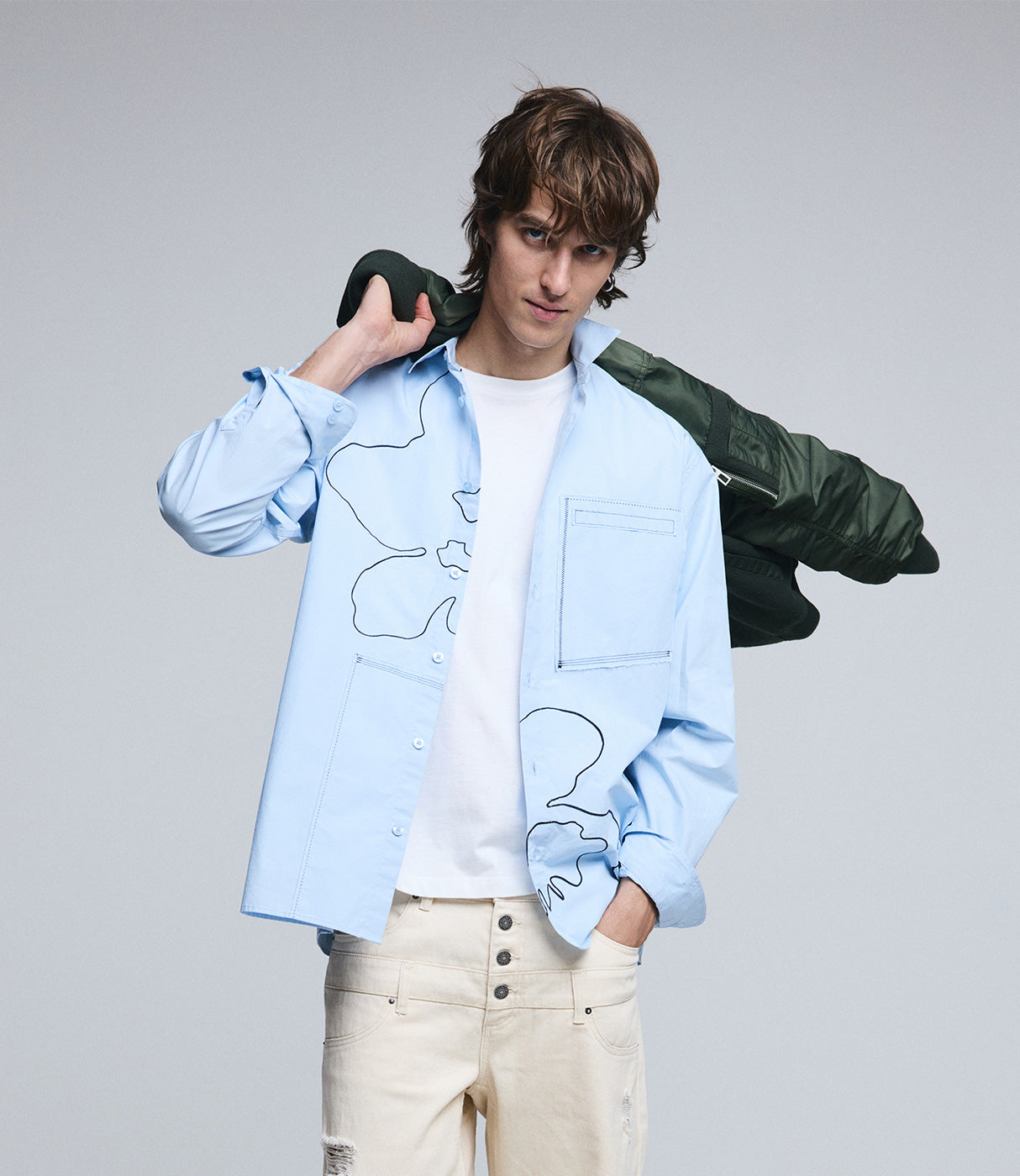גבר צעיר עם חולצה מכופתרת כחולה בעיצוב קווי מתאר, ג'ינס בז' ומעיל ירוק על הכתף