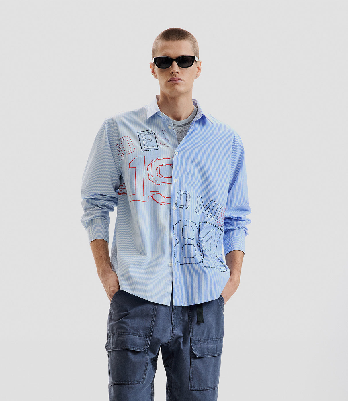 גבר עם חולצה מכופתרת תכלת עם רקמת מספרים, מכנסיים כחולים ומשקפי שמש, אופנת רחוב לגבר