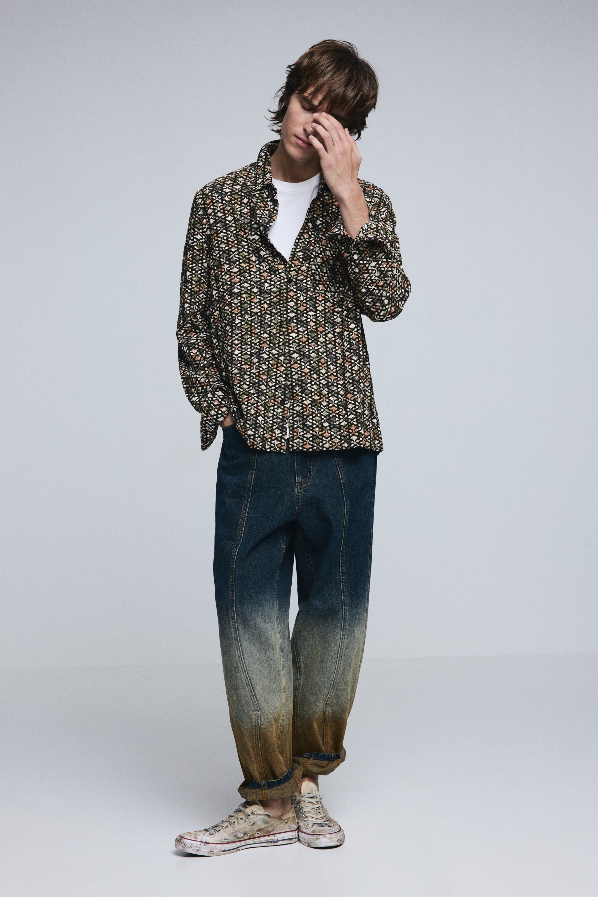 גבר לובש חולצה מודפסת צבעונית, חולצת טי לבנה, וג'ינס בגזרת אוברסייז בגווני דהייה, נעלי סניקרס