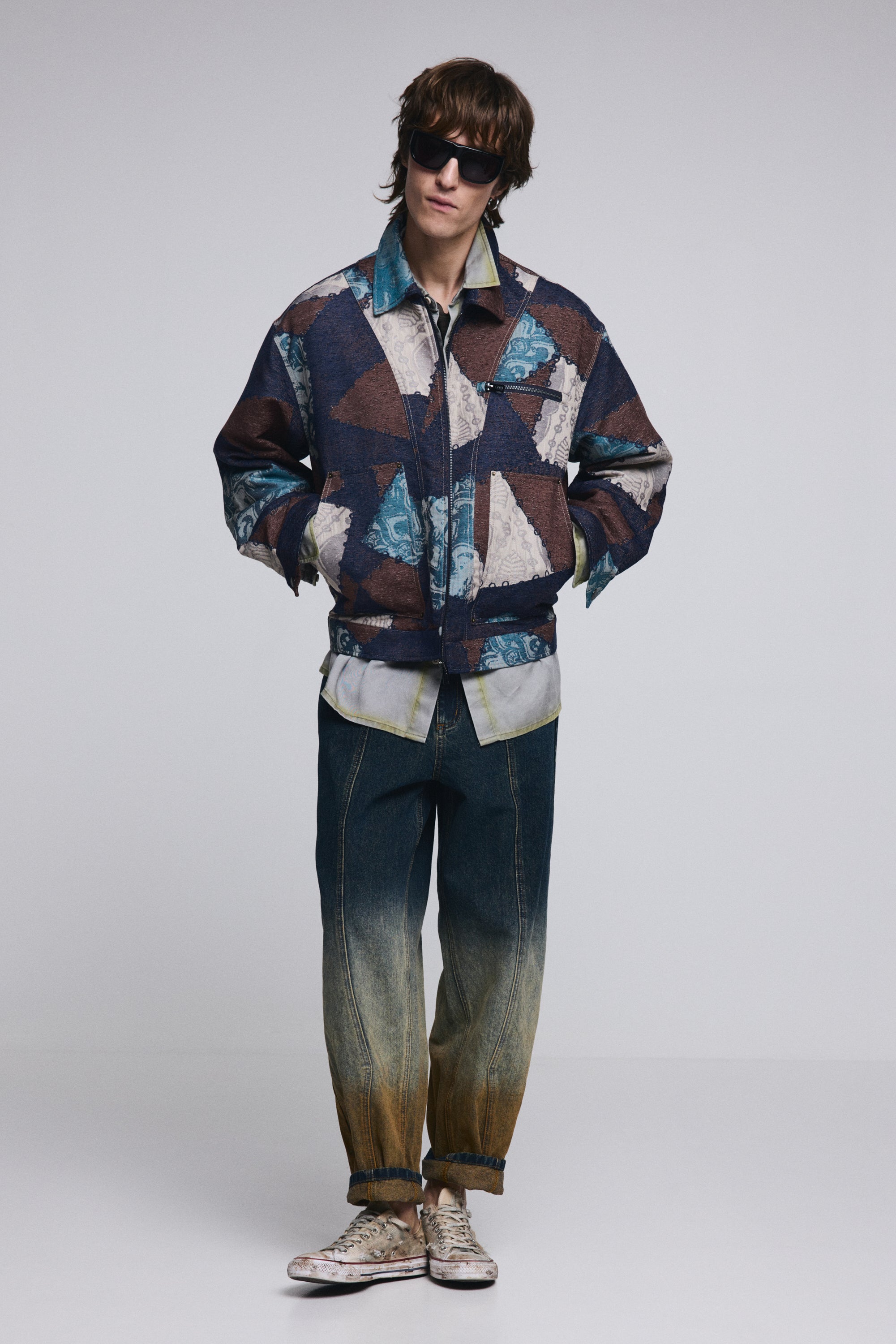 דגם גברי עם ז'קט פאטצ'ים צבעוני, ג'ינס דהוי ונעלי סניקרס טרנדיות, על רקע ניטרלי