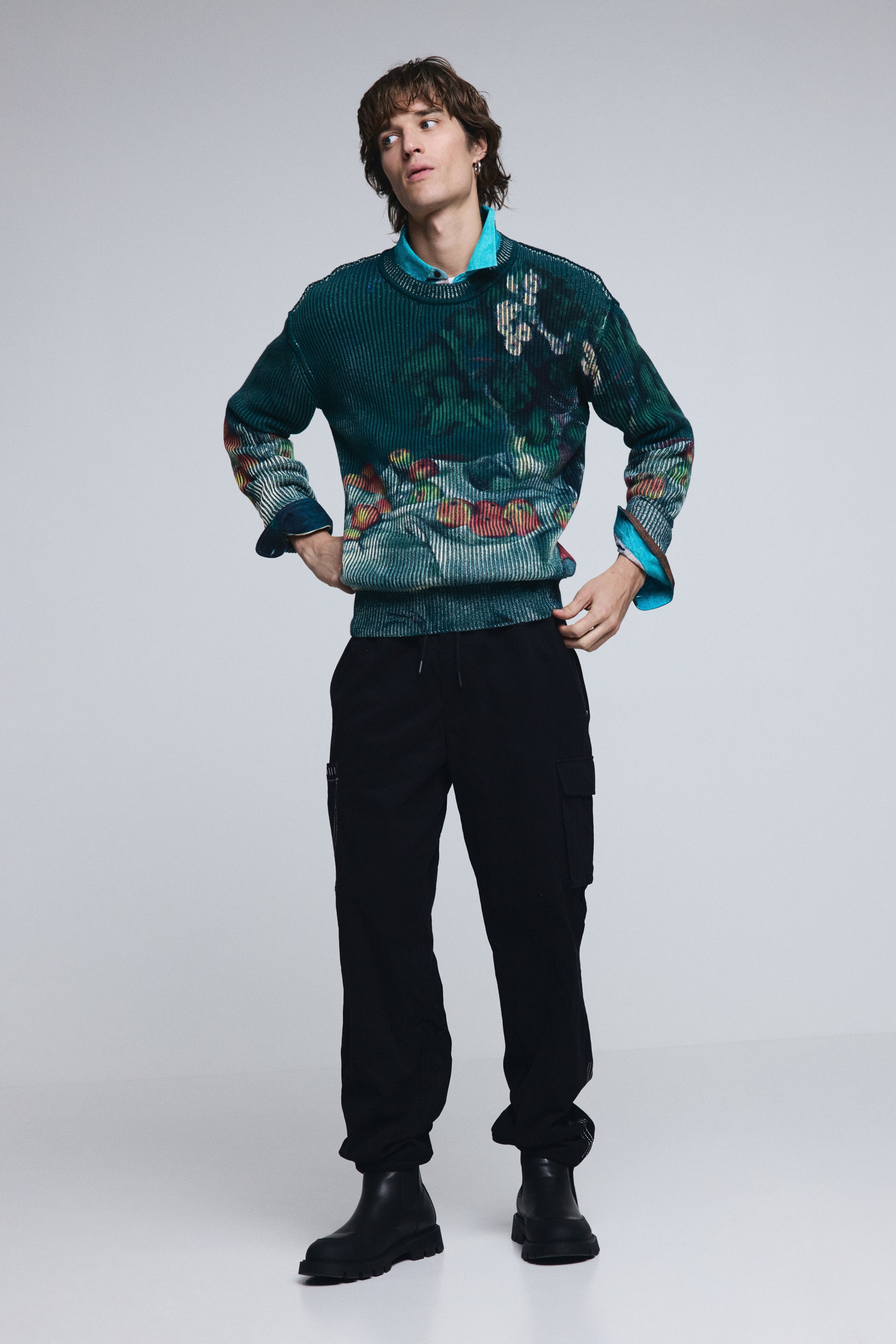 דוגמן לובש סוודר ירוק מודפס, חולצה כחולה ומכנסיים שחורים עם כיסים ומגפיים שחורים, אופנת גברים חורף