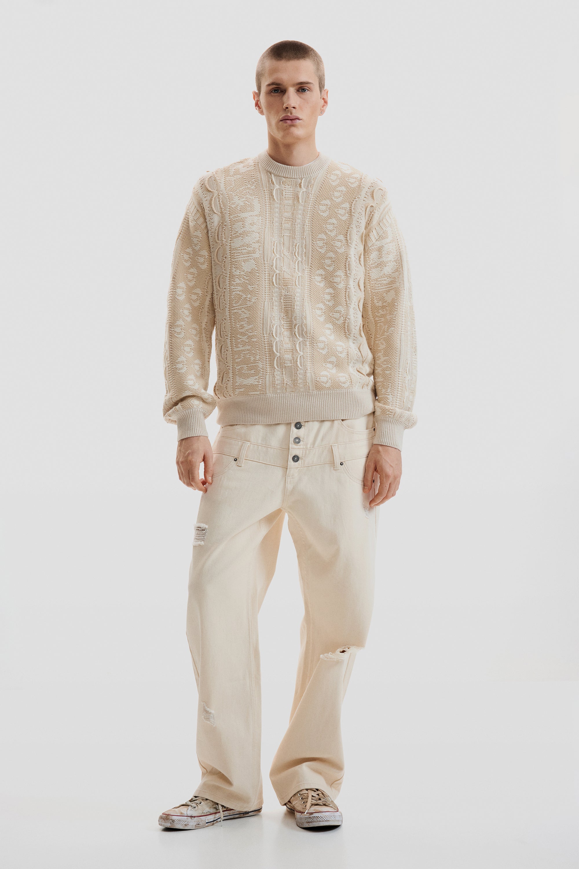 דגם גבר לובש סוודר בצבע בז' עם דוגמה סרוגה ומכנסי ג'ינס בז' רחבים עם קרעים אופנתיים באווירה מודרנית