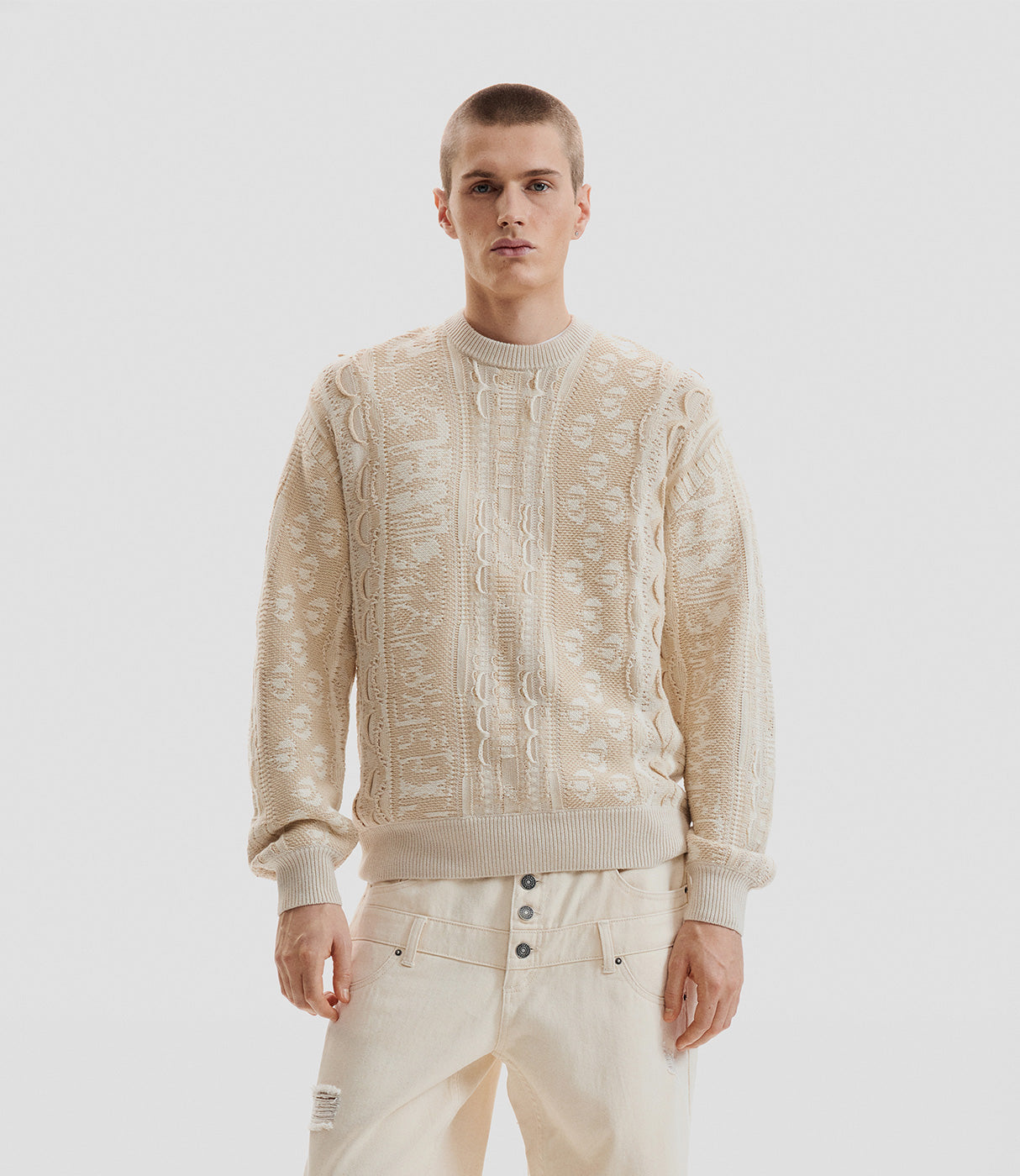 גבר מדגמן סוודר בז' סרוג עם דוגמה ייחודית ומכנסי ג'ינס בהירים, אופנת גברים אלגנטית