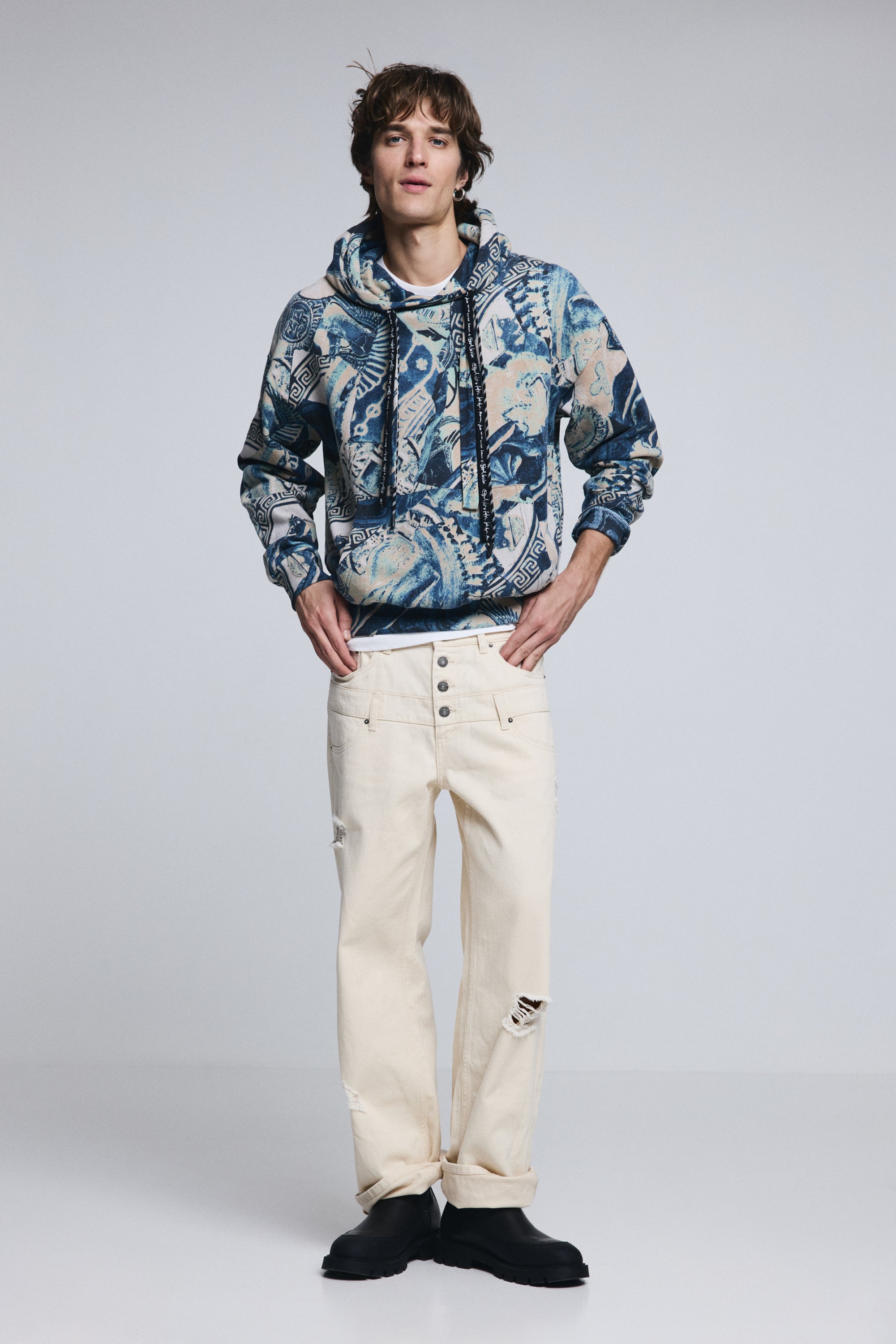 גבר מדגמן סווטשירט קפוצ'ון עם דוגמה כחולה ובז', מכנסיים לבנים קרועים ונעלי בוקרים שחורות