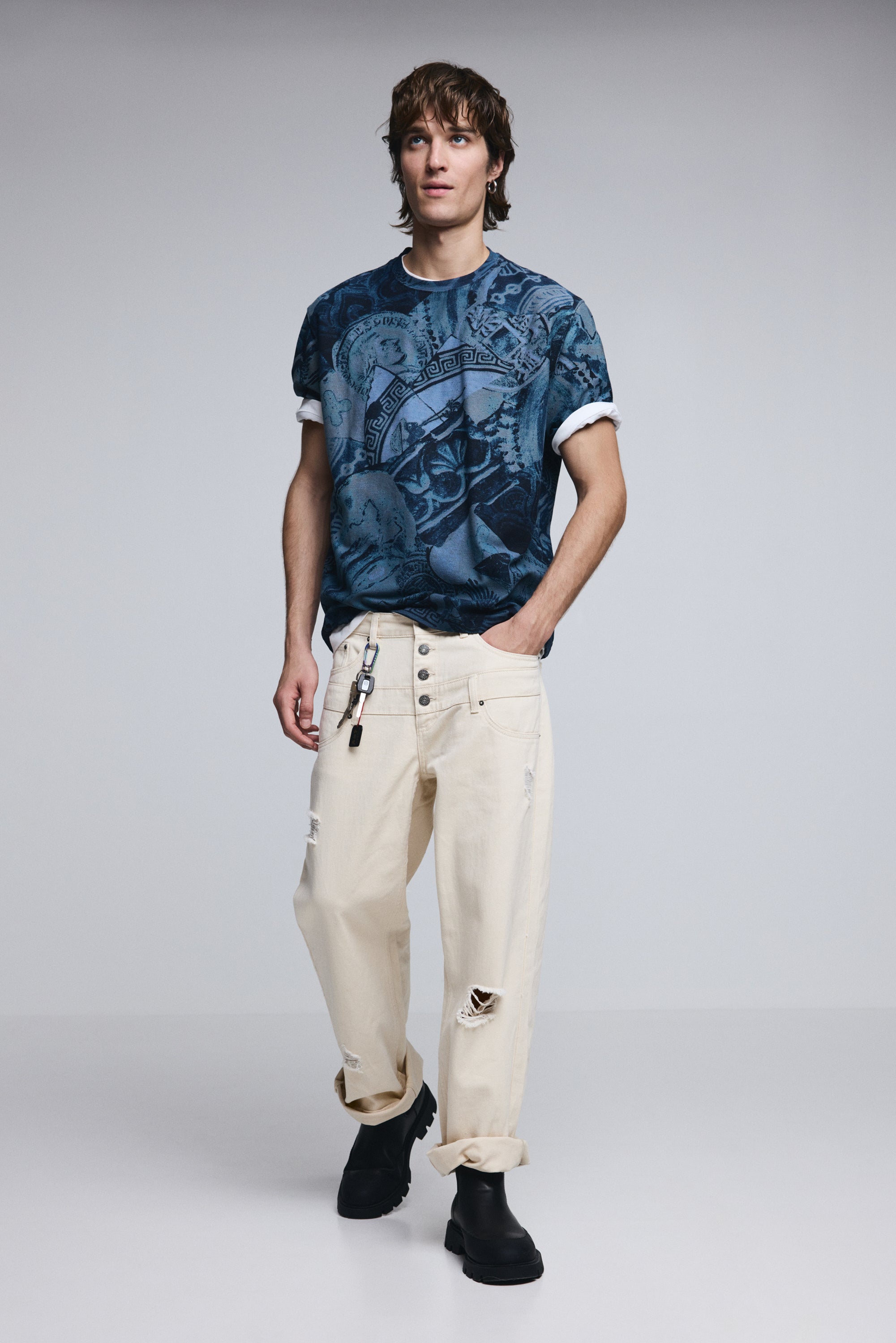 דוגמן לובש חולצת טריקו כחולה עם הדפס ודג'ינס בז' קרוע, מגפי עור שחורים, אופנת גברים עדכנית