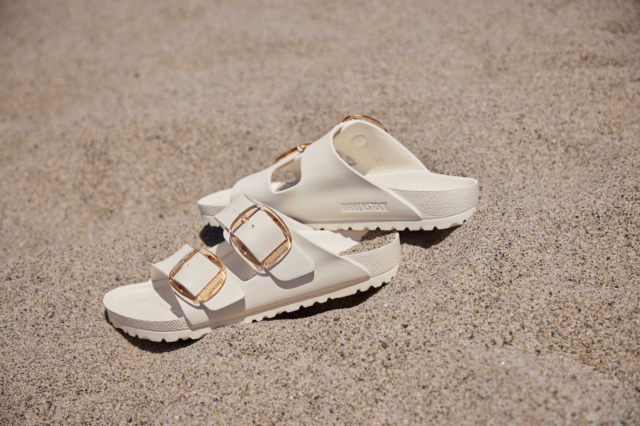 סנדלי בירקנשטוק לבנים עם אבזמי זהב על חול, נעליים ממותגי ייבוא רשמי CHOZEN
