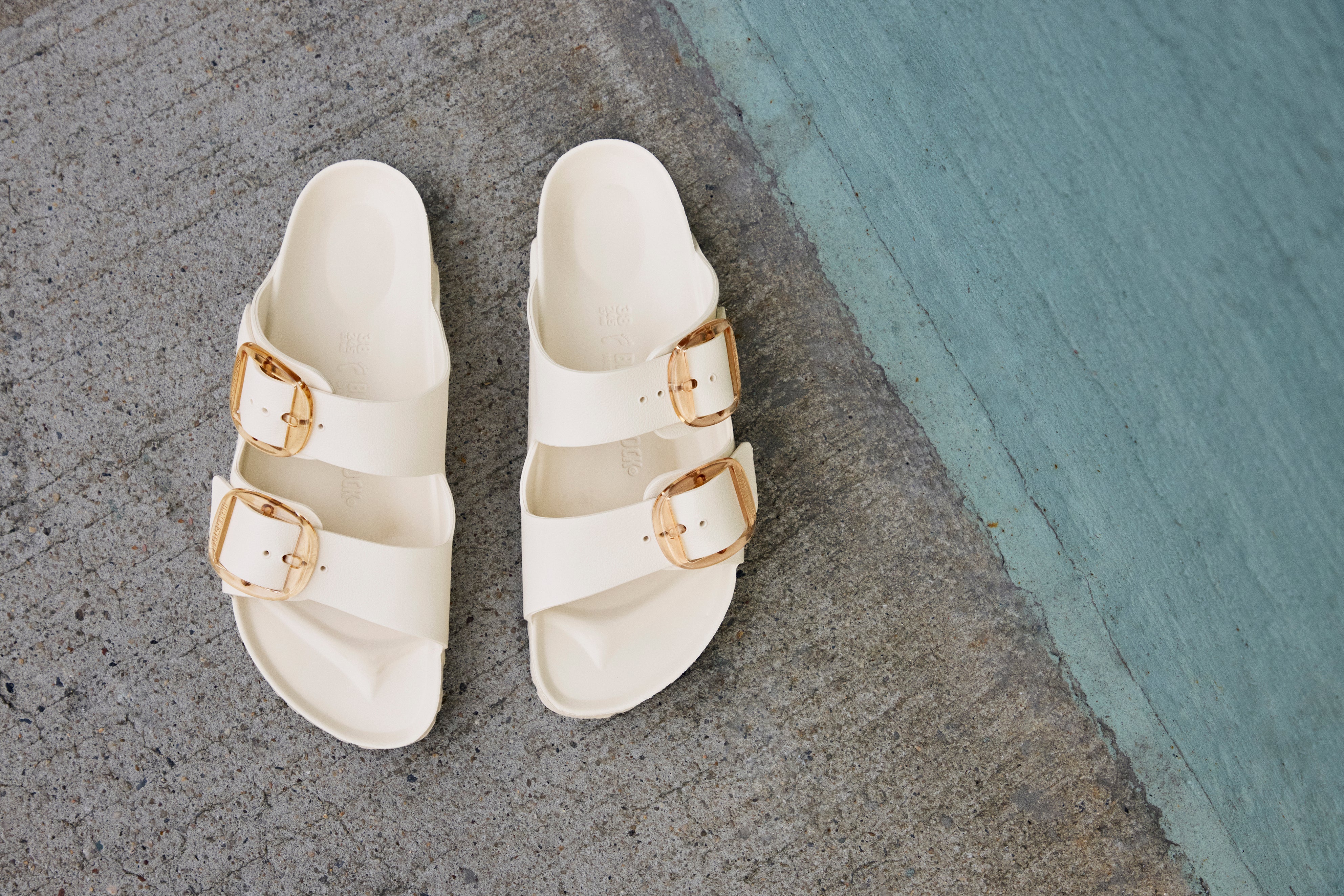 כפכפי נשים לבנים עם אבזמים מוזהבים על רצפת בטון, מותג CHOZEN אלפא