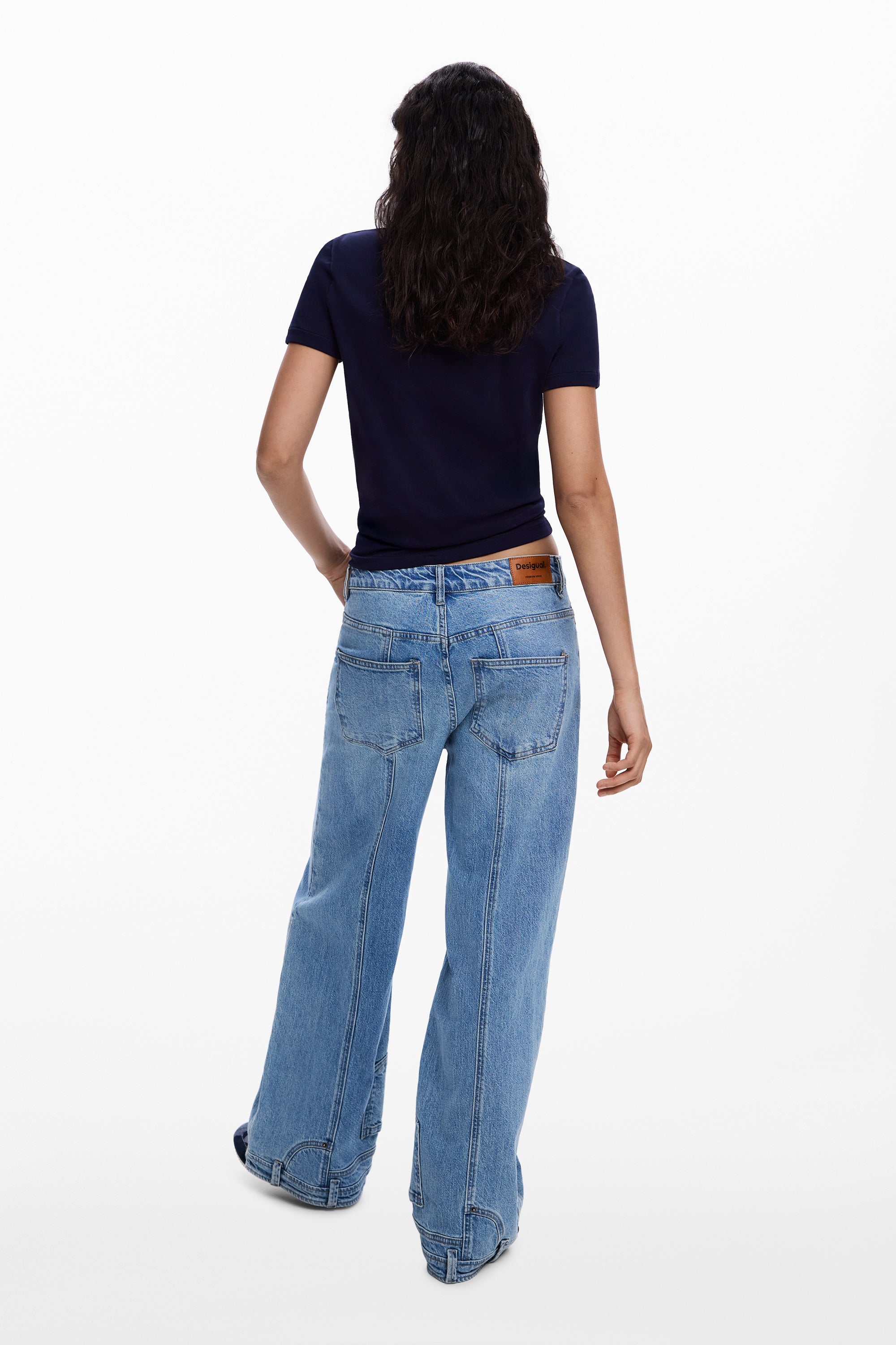 אישה עם שיער כהה לובשת חולצת טי כחולה ומכנסי ג'ינס רחבים בגזרה גבוהה, מבט מאחור, רקע לבן