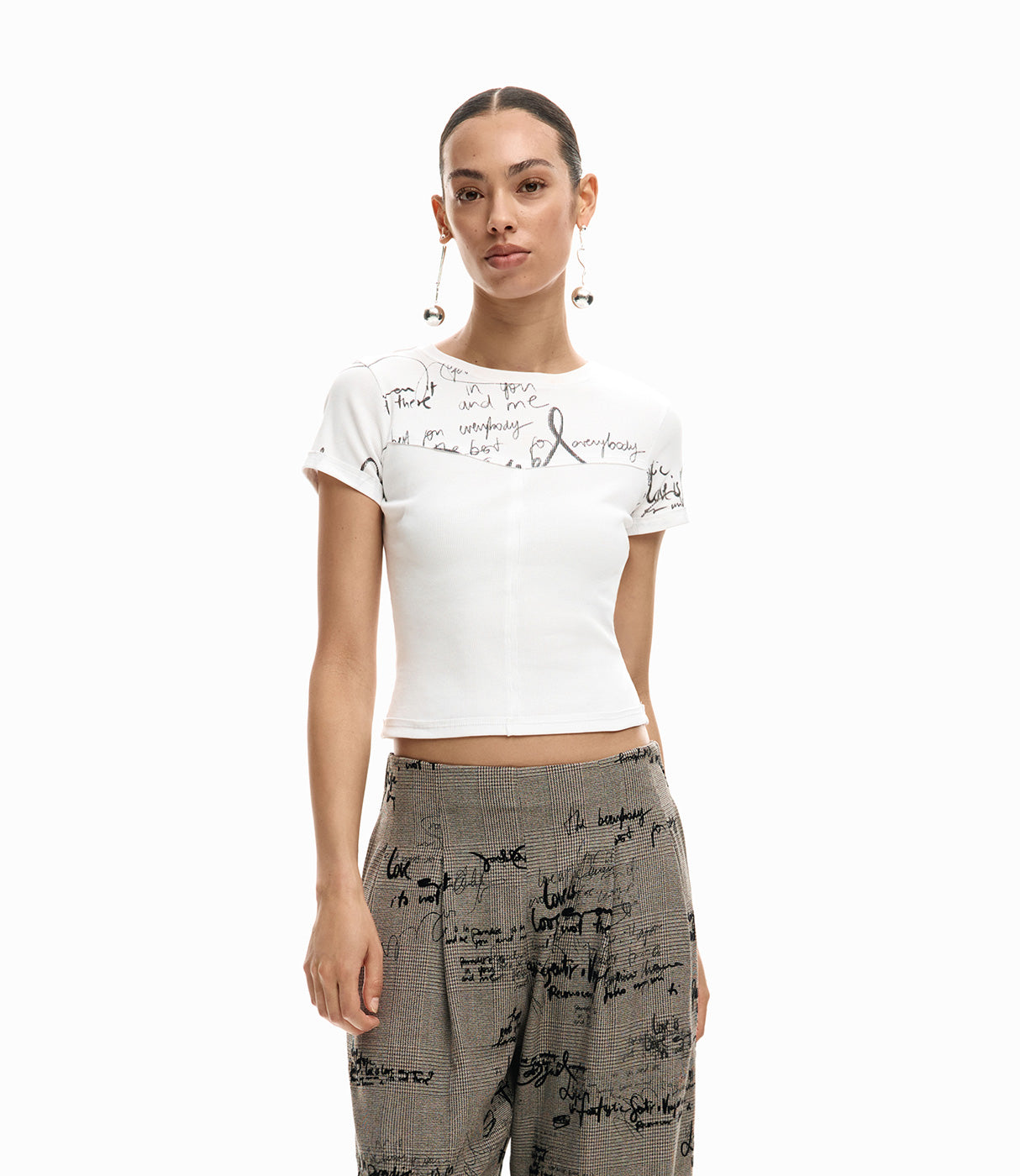 אישה לובשת חולצה לבנה קצרה עם הדפס טקסט שחור ומכנסיים אפורים מודפסים, צילום אופנה על רקע לבן