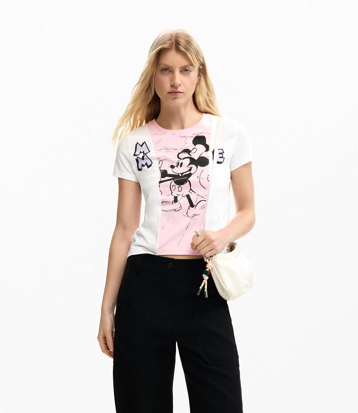 אישה לובשת חולצת טי קצרה לבנה עם הדפס דמות מצוירת ורקע ורוד, מכנסיים שחורים ותיק קטן