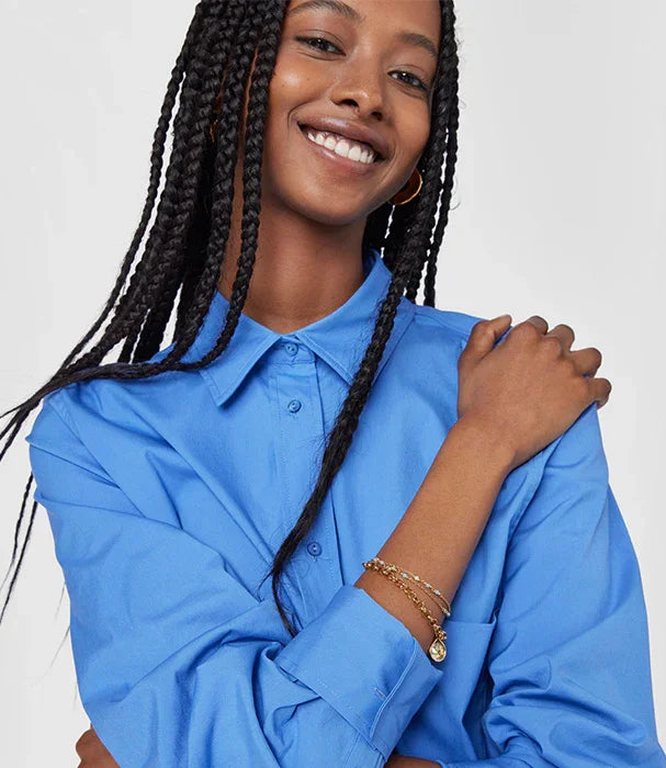 אישה מחייכת בחולצה כחולה, עונדת צמידי זהב, מראה אופנה אלגנטית מבית CHOZEN