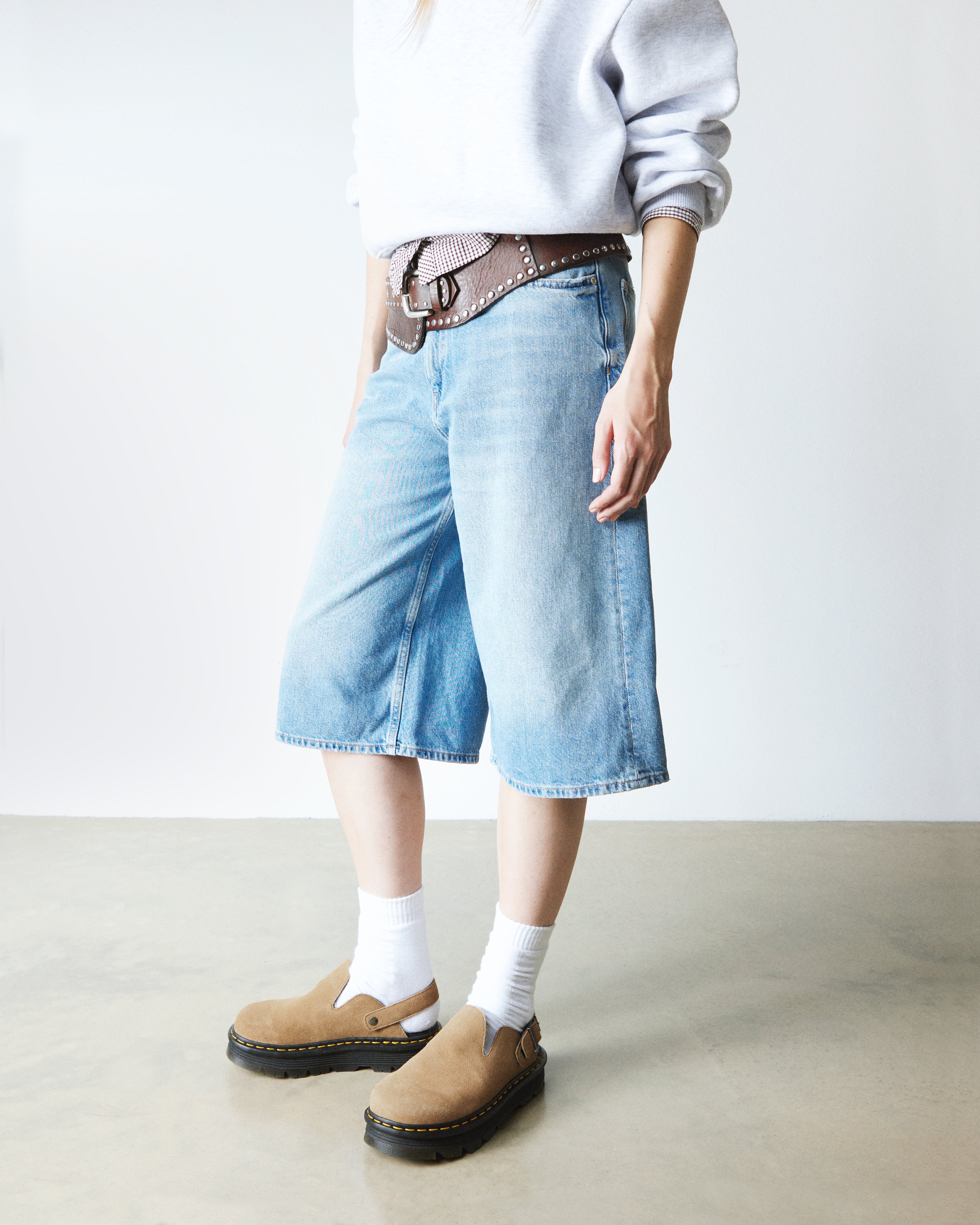 אישה בלבוש קז'ואל עם מכנסי ג'ינס קצרים, חגורת ניטים חומה ונעלי מוקסין חומות על רקע לבן