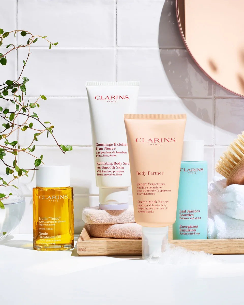 מוצרי טיפוח של קלרינס על מדף בחדר אמבטיה, מוצגים ליד צמח ומגבת, מותגים מובילים מבית אלפא