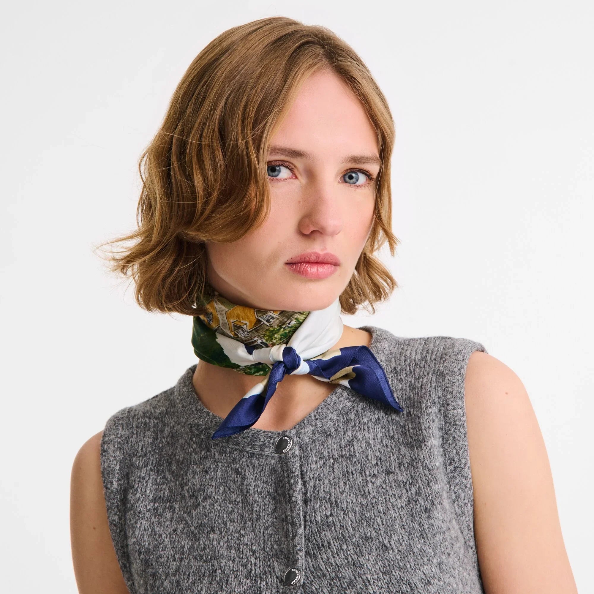 אישה עם צעיף משי מעוצב בגווני כחול ולבן מסביב לצוואר, לובשת אפודה אפורה, מותג CHOZEN