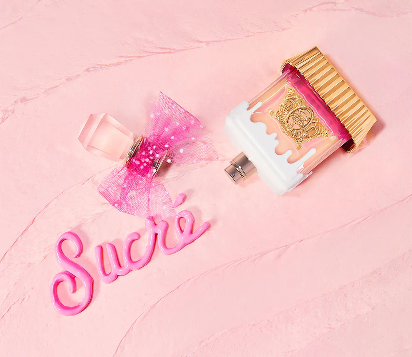 בקבוק בושם מעוצב בגווני ורוד וזהב עם פקק יהלום וסרט פפיון, רקע ורוד ומילה Sucré