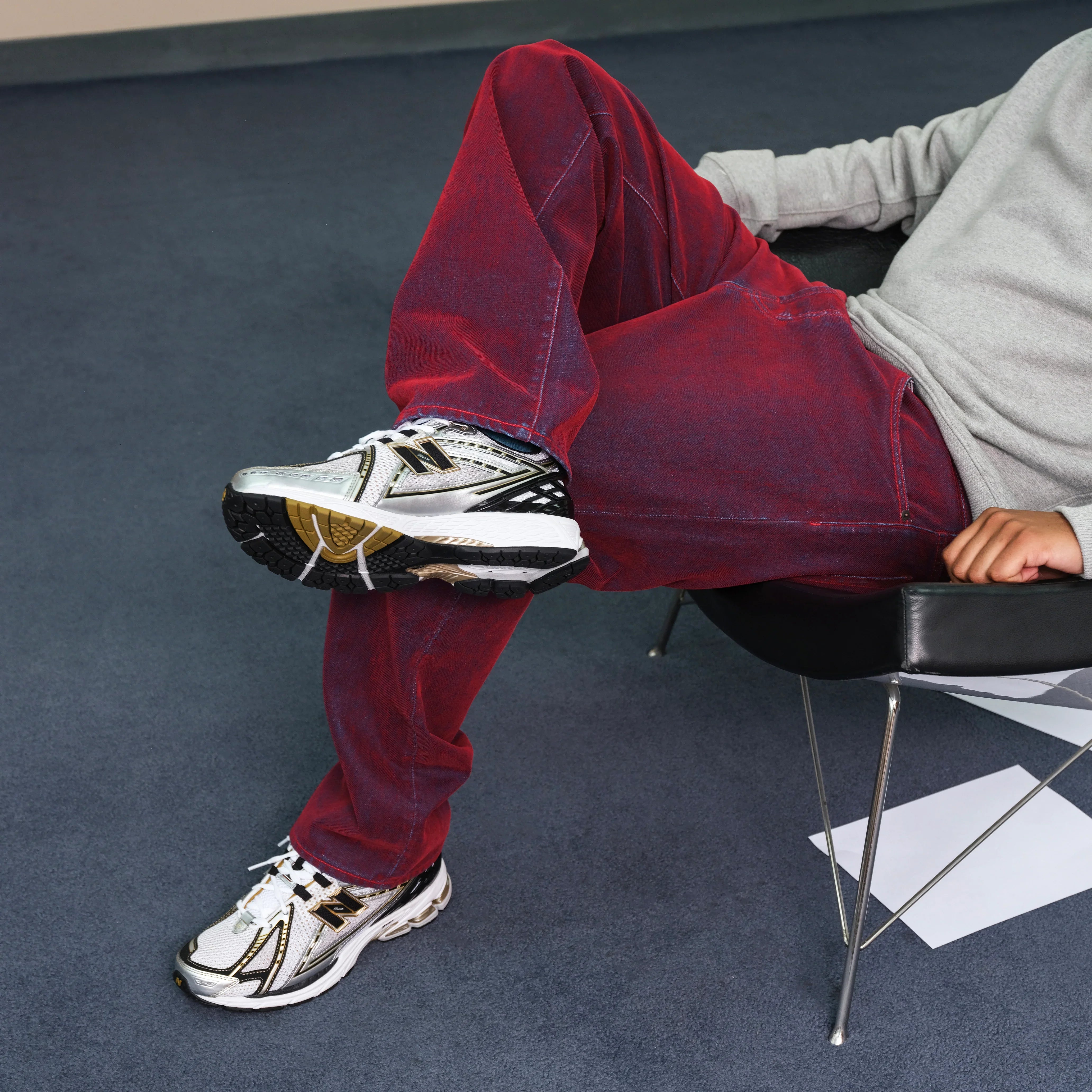 אדם יושב על כיסא עם ג'ינס אדום, נעלי ניו באלאנס כסופות וחולצה אפורה, CHOZEN אופנה יוקרתית